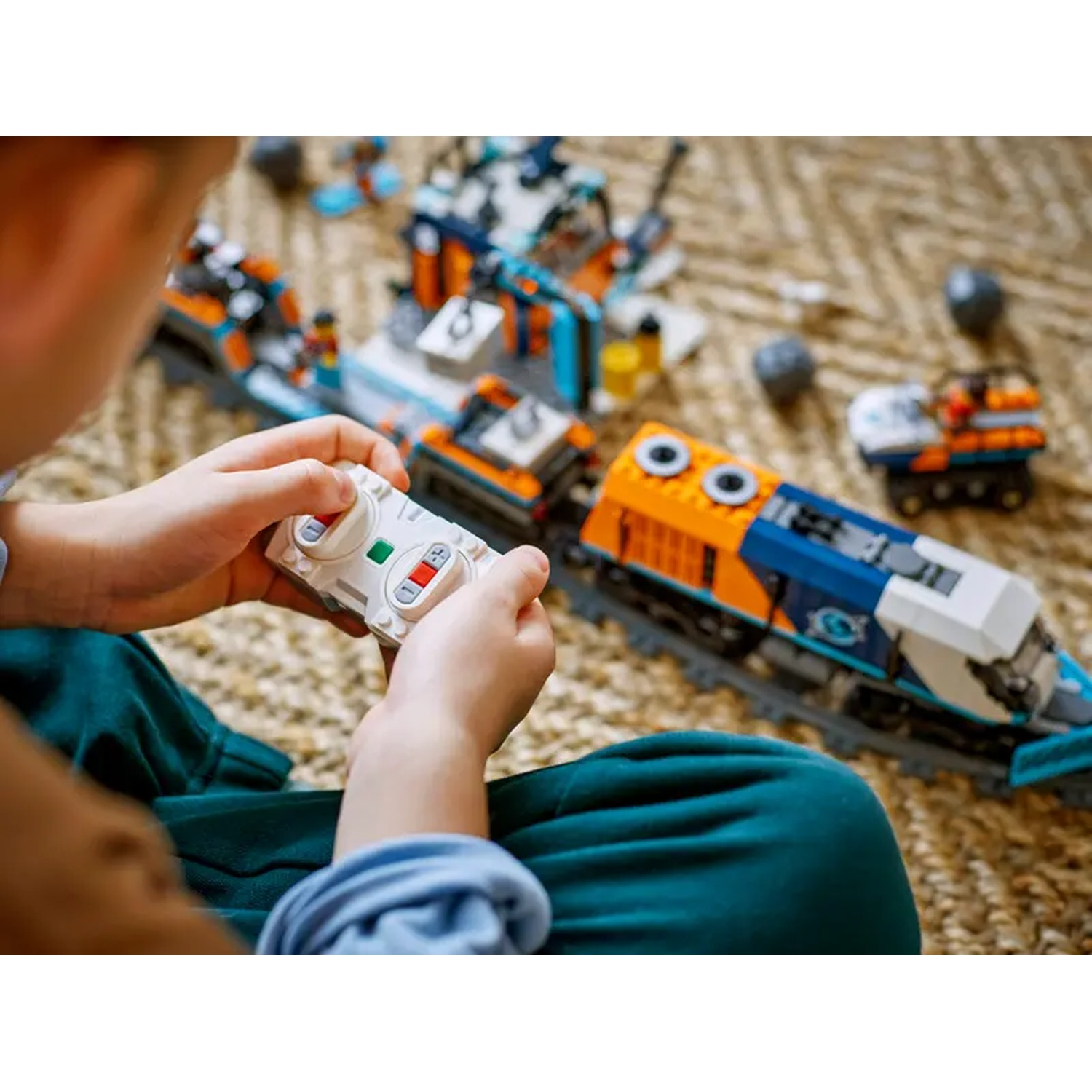 A child holds a white remote, operating a toy train on a rug. The train is blue and orange. Other toy parts are scattered.