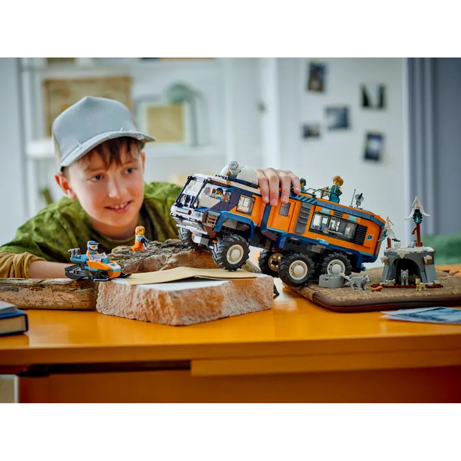 A boy smiles, holding a Lego truck. Other Lego sets include a snowmobile, a cave, and small figures, all on a yellow table.
