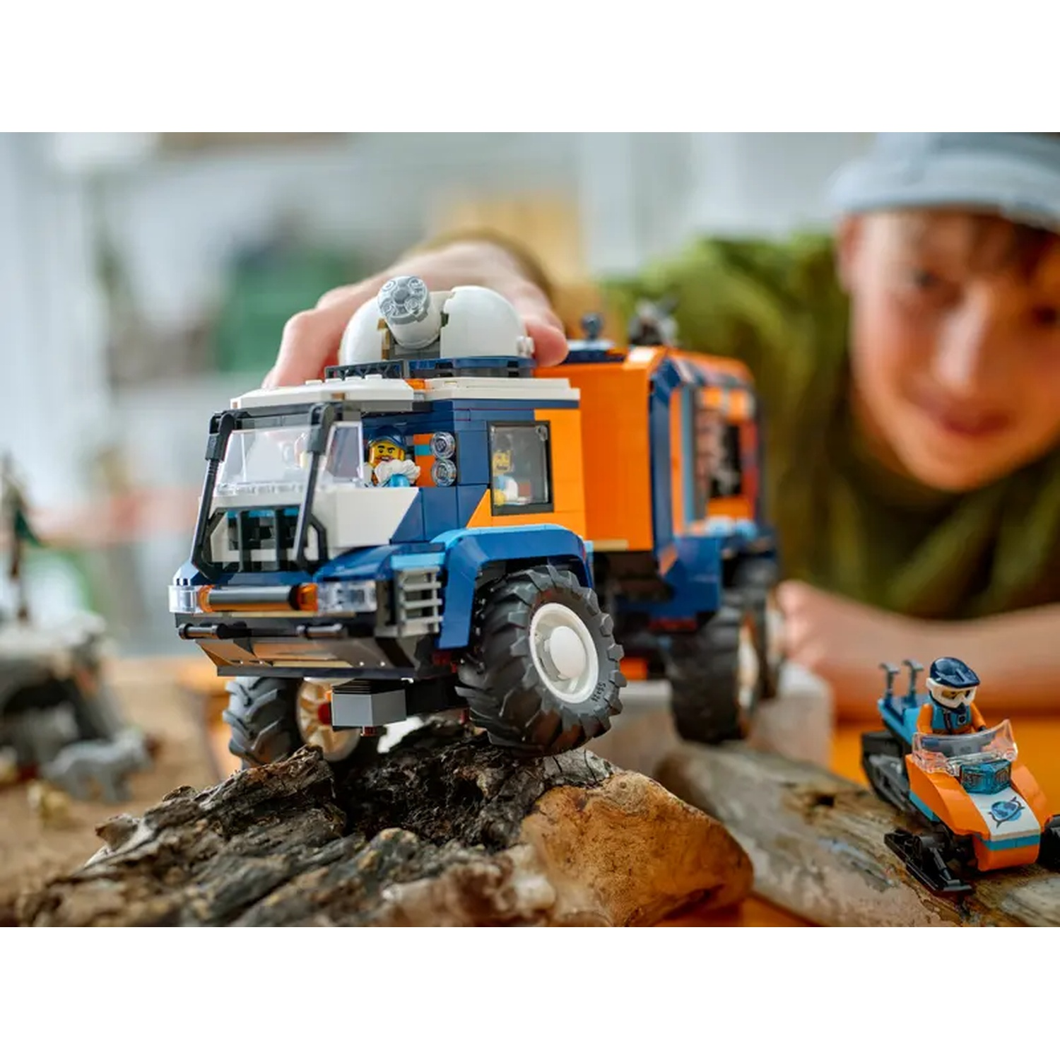 A child plays with a Lego truck on a wooden surface. The truck is blue and orange. A snowmobile is also present.