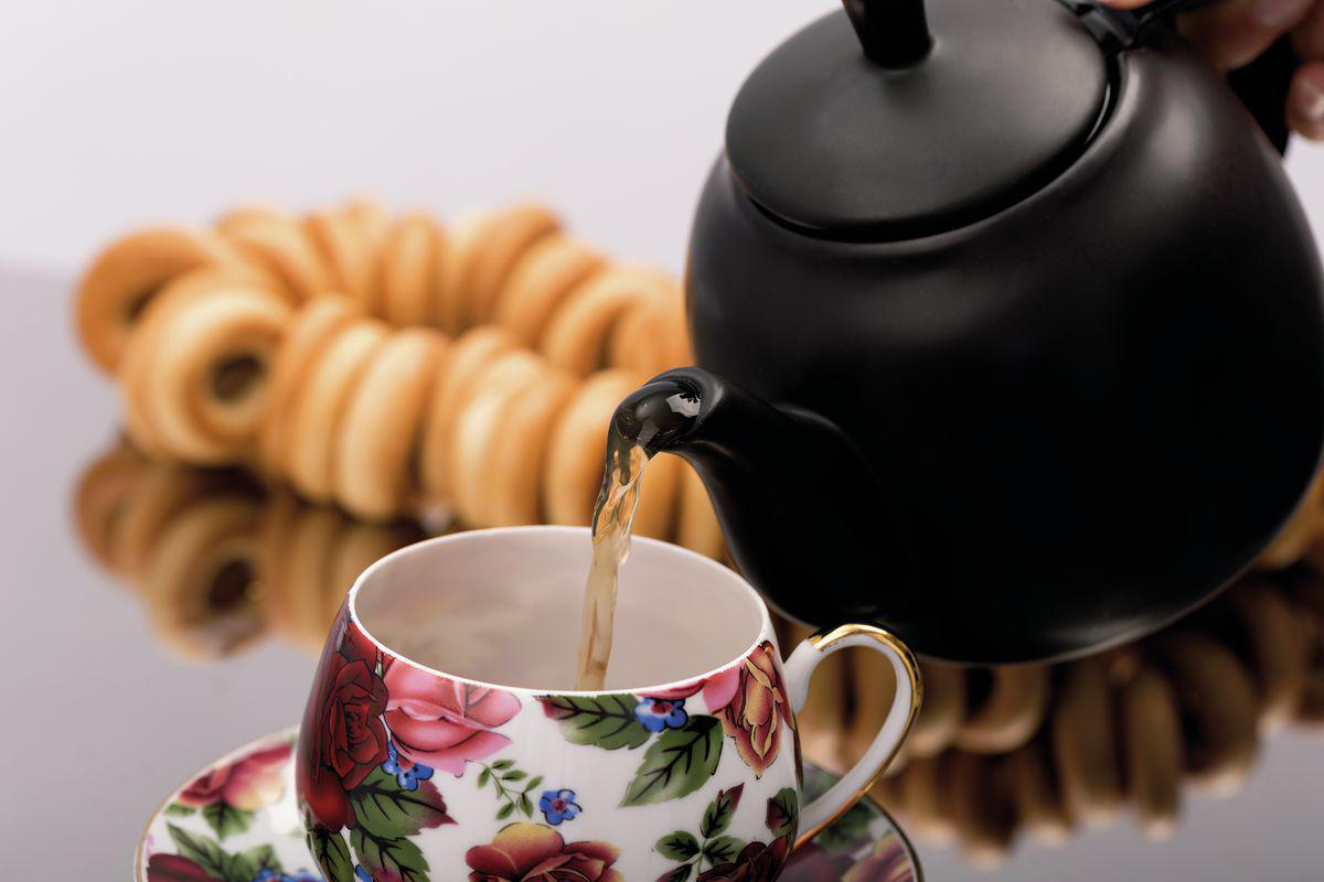 Ein schwarzer Teekessel gießt Flüssigkeit in eine Blumetasse. Bagels sind im Hintergrund.