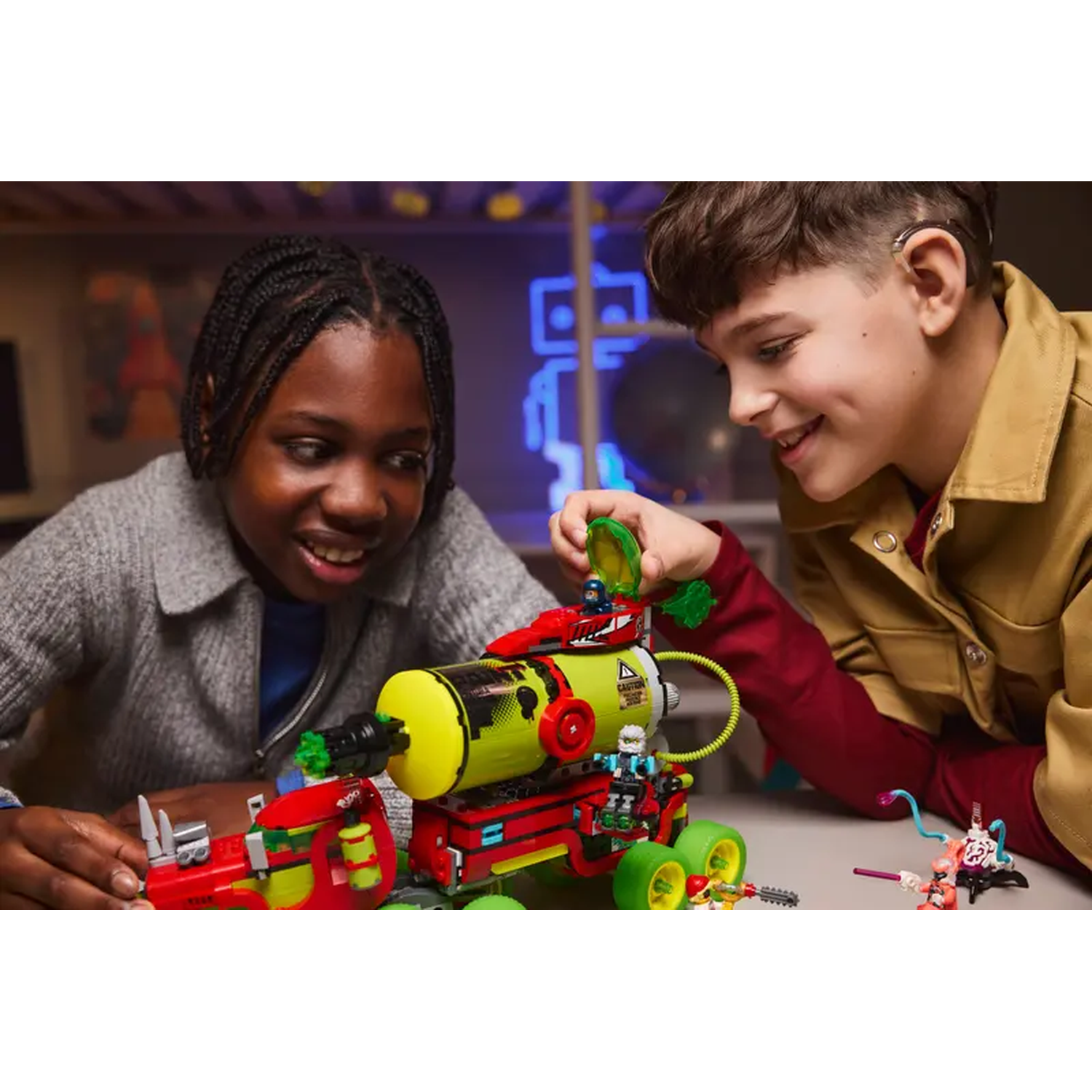 Two children play with a colorful toy vehicle. The toy is red, yellow, and green. They are smiling.