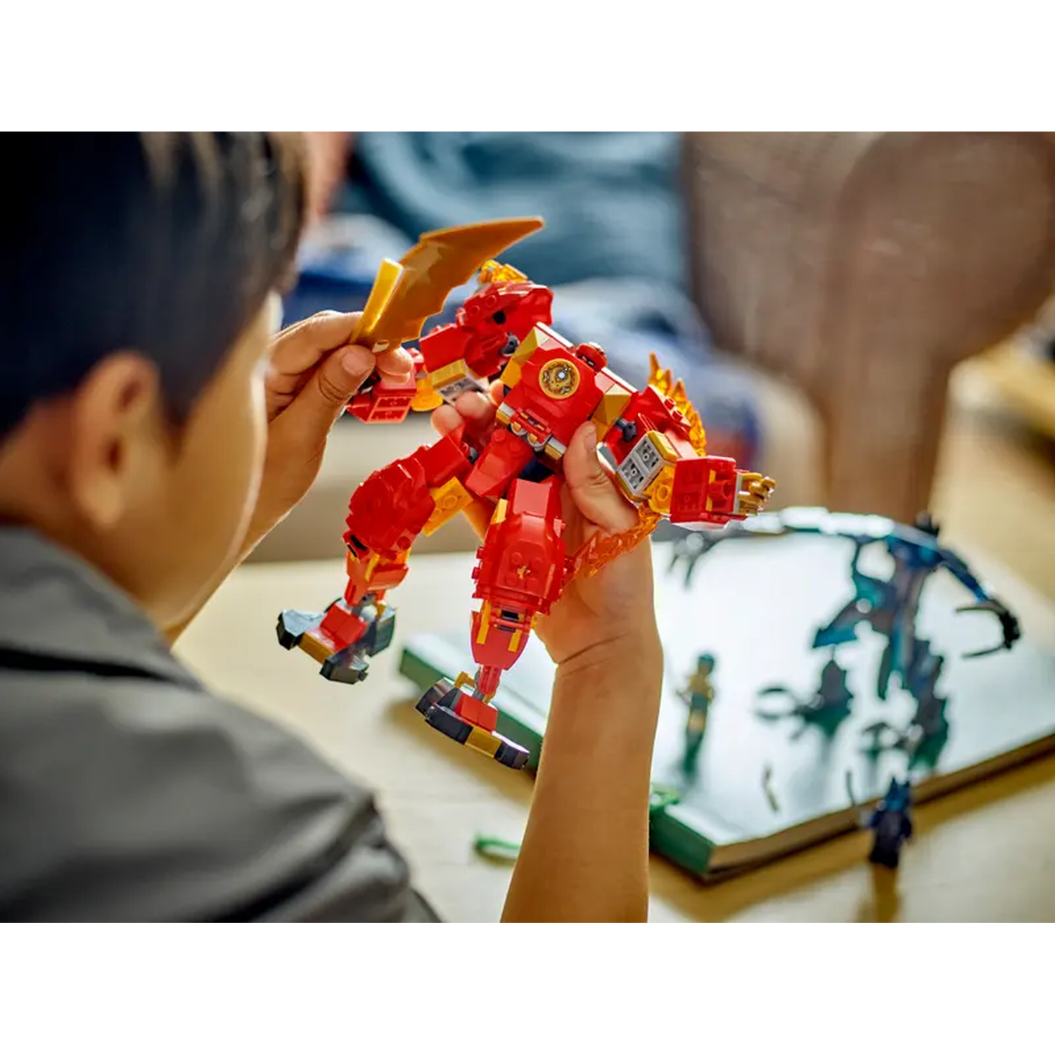 Een kind houdt een rood-gele Lego-robot vast met een zwaard. Andere Lego-figuren en een boek liggen op een tafel.