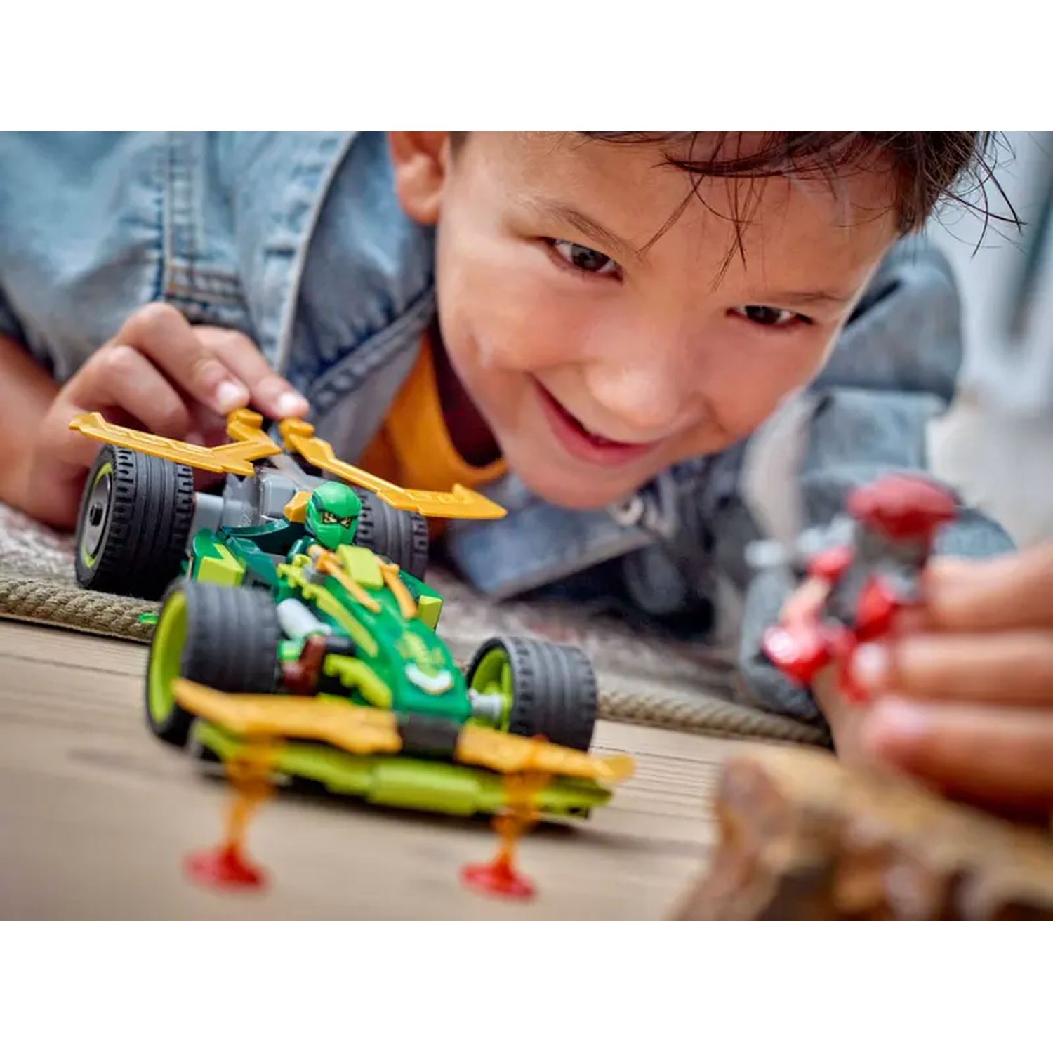 A boy smiles, playing with a green toy car. The car has yellow wings and is on a wooden surface. Another red toy is near.