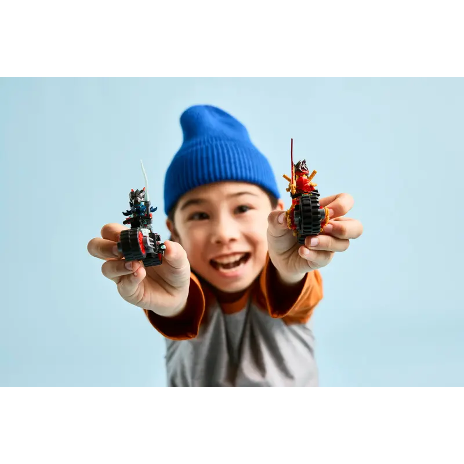 A smiling boy holds two toy vehicles. One is black and red, the other is red and gold. He wears a blue hat.