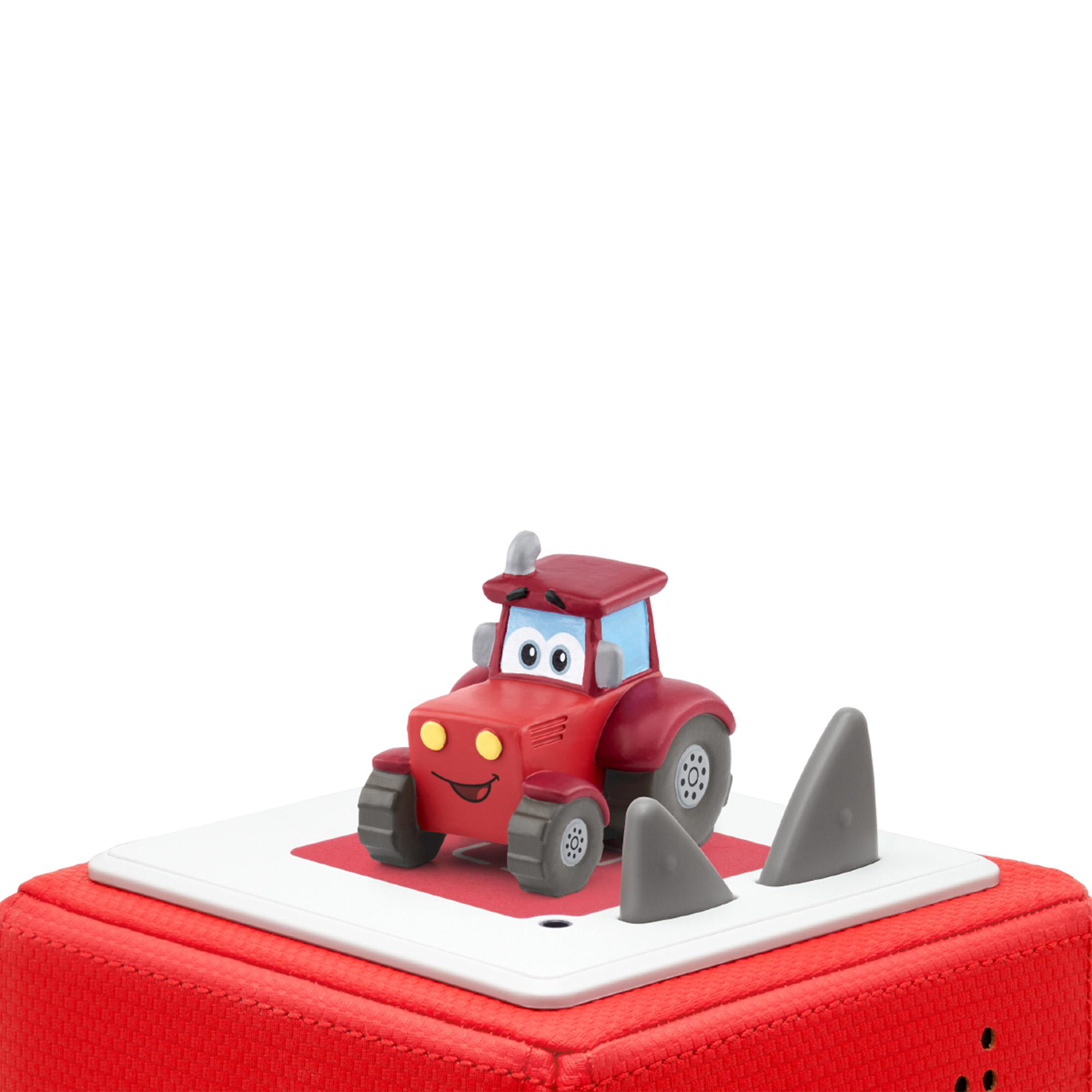 Ein roter Spielzeugtraktor mit lächelndem Gesicht auf einem roten Block. Graue Haifischflossen daneben. Weißer Hintergrund.