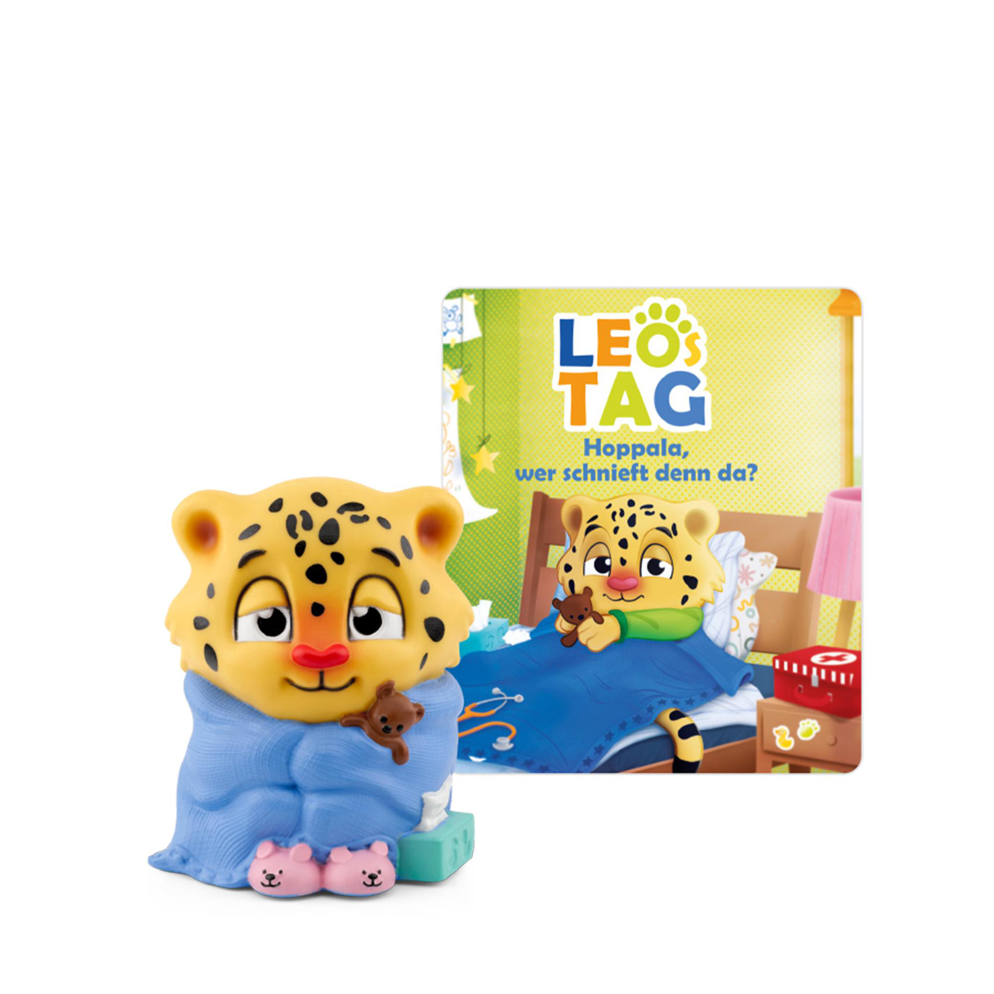Ein Spielzeugleopard, eingewickelt in eine blaue Decke, hält einen Teddybären. Ein Buch zeigt eine ähnliche Szene. Rosa Bärenhausschuhe sind sichtbar.