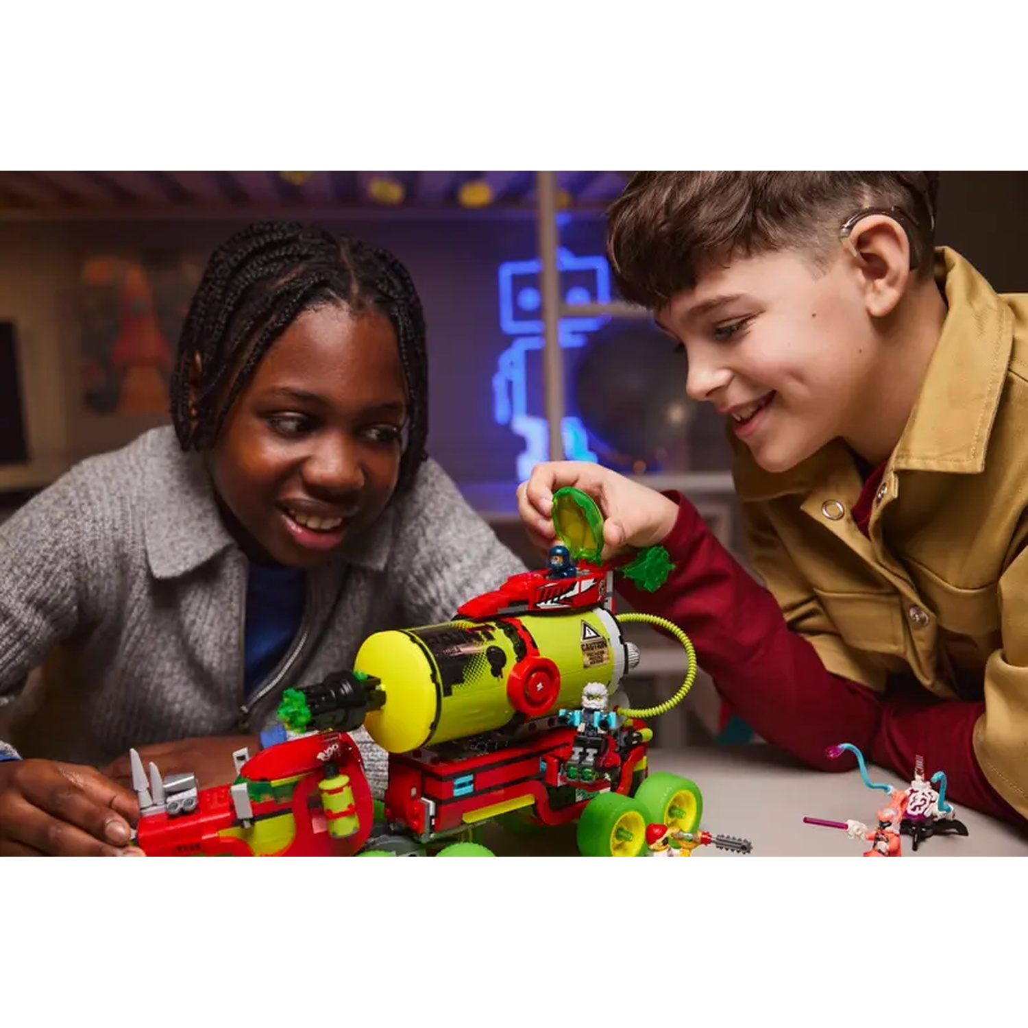 Twee kinderen glimlachen terwijl ze met een kleurrijk speelgoedvoertuig spelen. Het voertuig is rood, groen en geel, met een kleine figuur erop.