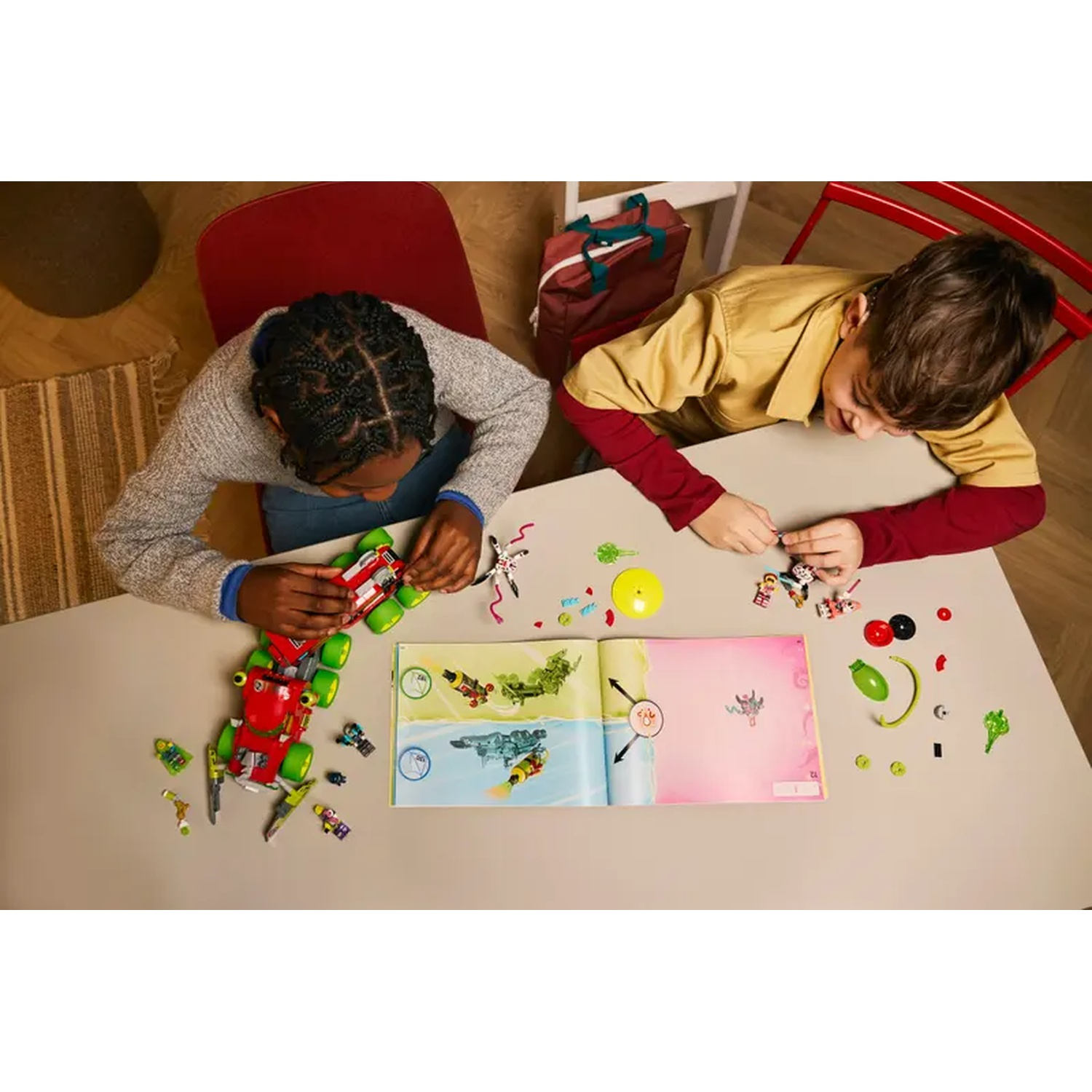 Twee kinderen zetten een speelgoedauto en figuren in elkaar op een tafel. Een boek ligt open. Verschillende onderdelen en accessoires liggen verspreid.