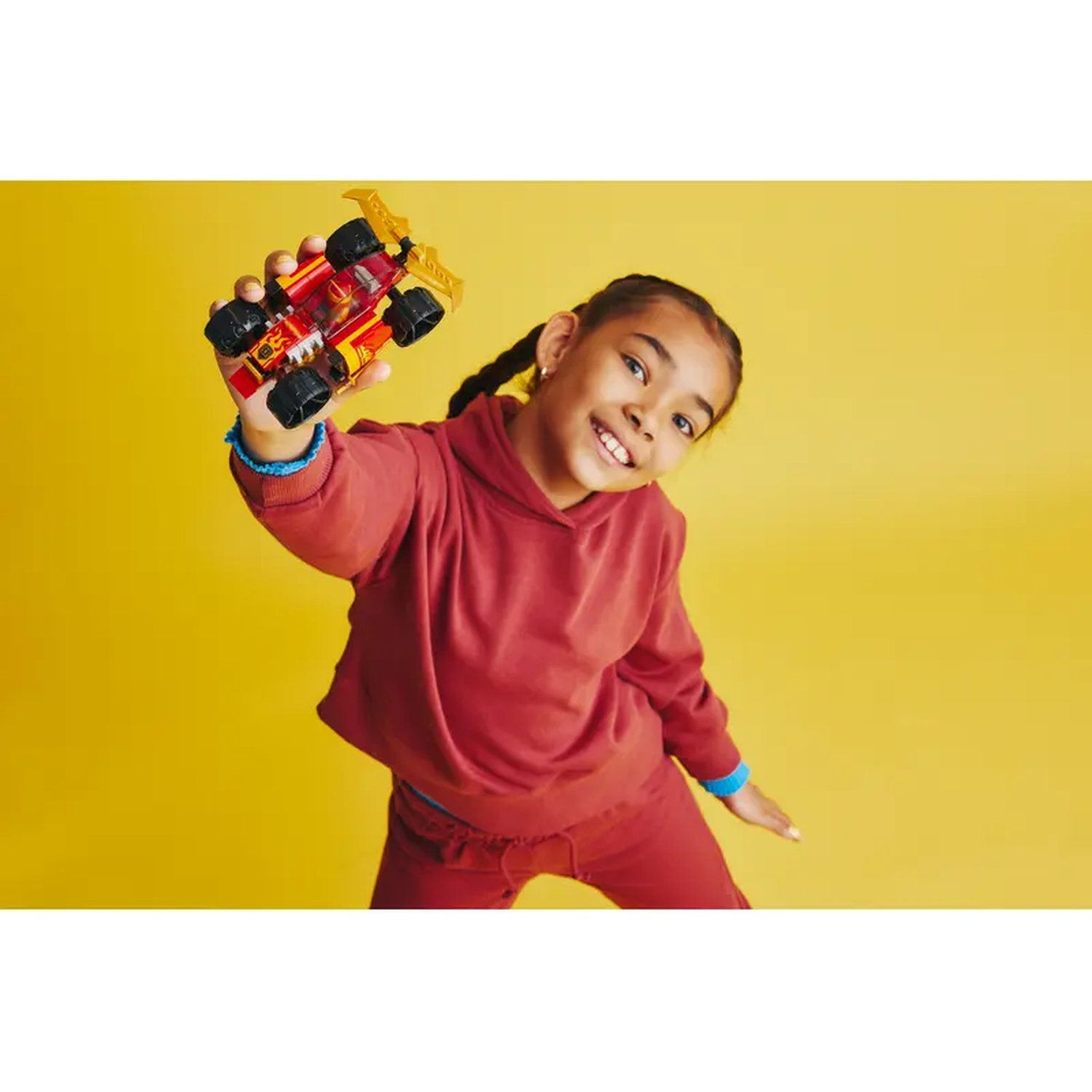 A girl smiles, holding a toy car. She wears a red sweatsuit against a yellow backdrop. The toy car is red, black, and gold.
