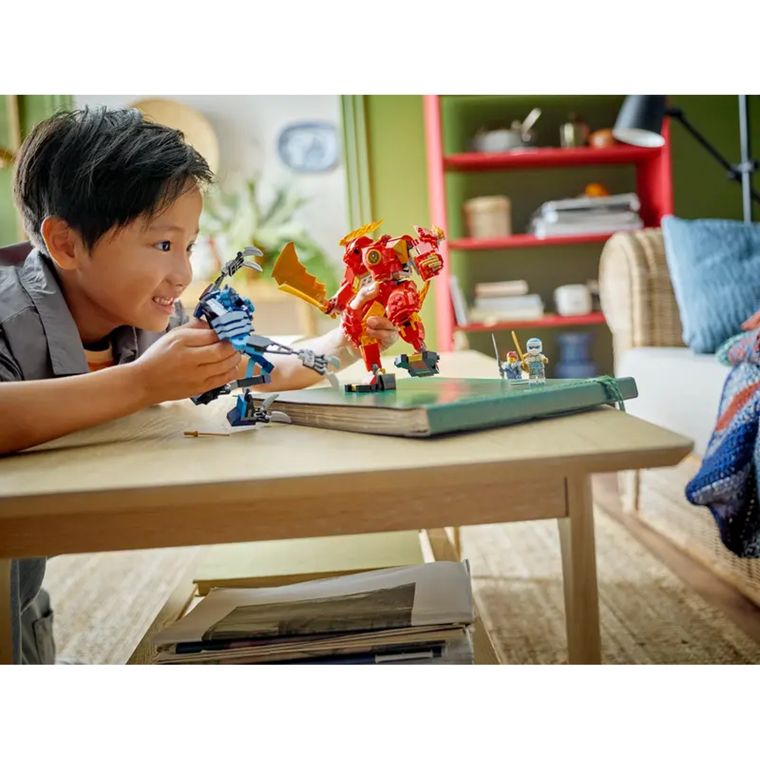Een jongen lacht, spelend met Lego-robots op een tafel. Een rode robot en een blauwe robot staan tegenover elkaar. Andere Lego-figuren op de achtergrond.