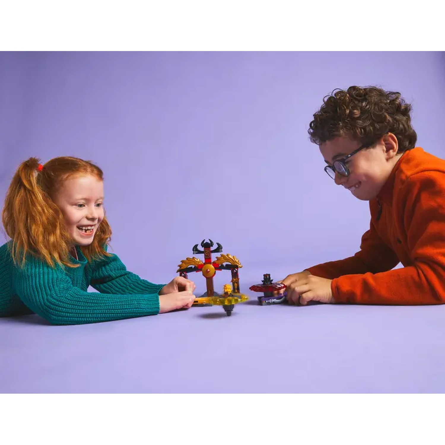 Two children playing with a toy set on a purple surface. The girl wears a green sweater, the boy a red one, both smiling.