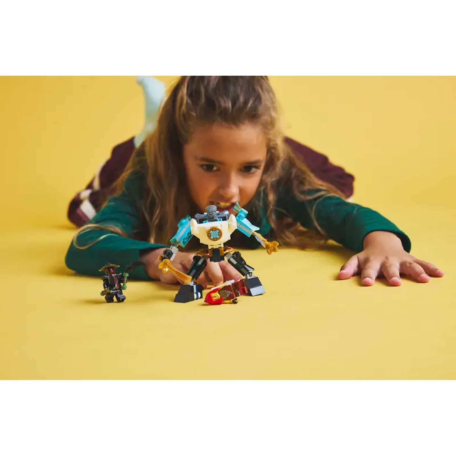 A girl lies on a yellow surface, playing with Lego robots. The robots are in the foreground, the girl in the background.