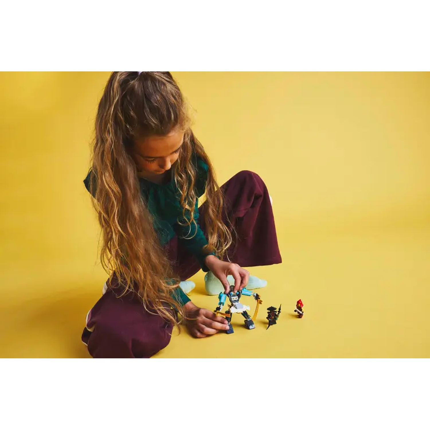 A girl plays with Lego figures on a yellow background. She is wearing green shirt and burgundy pants. The Lego figures are black and red.