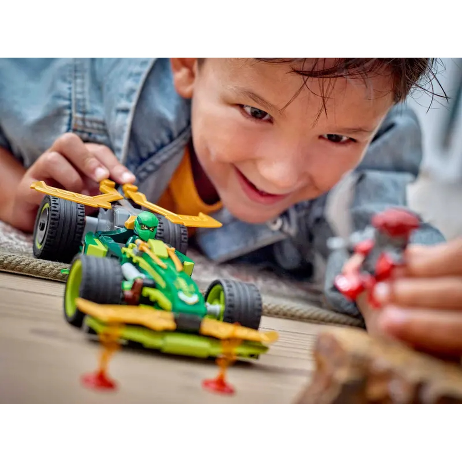 A smiling boy plays with a green and yellow toy car. A red toy is in his other hand. The background is blurred.