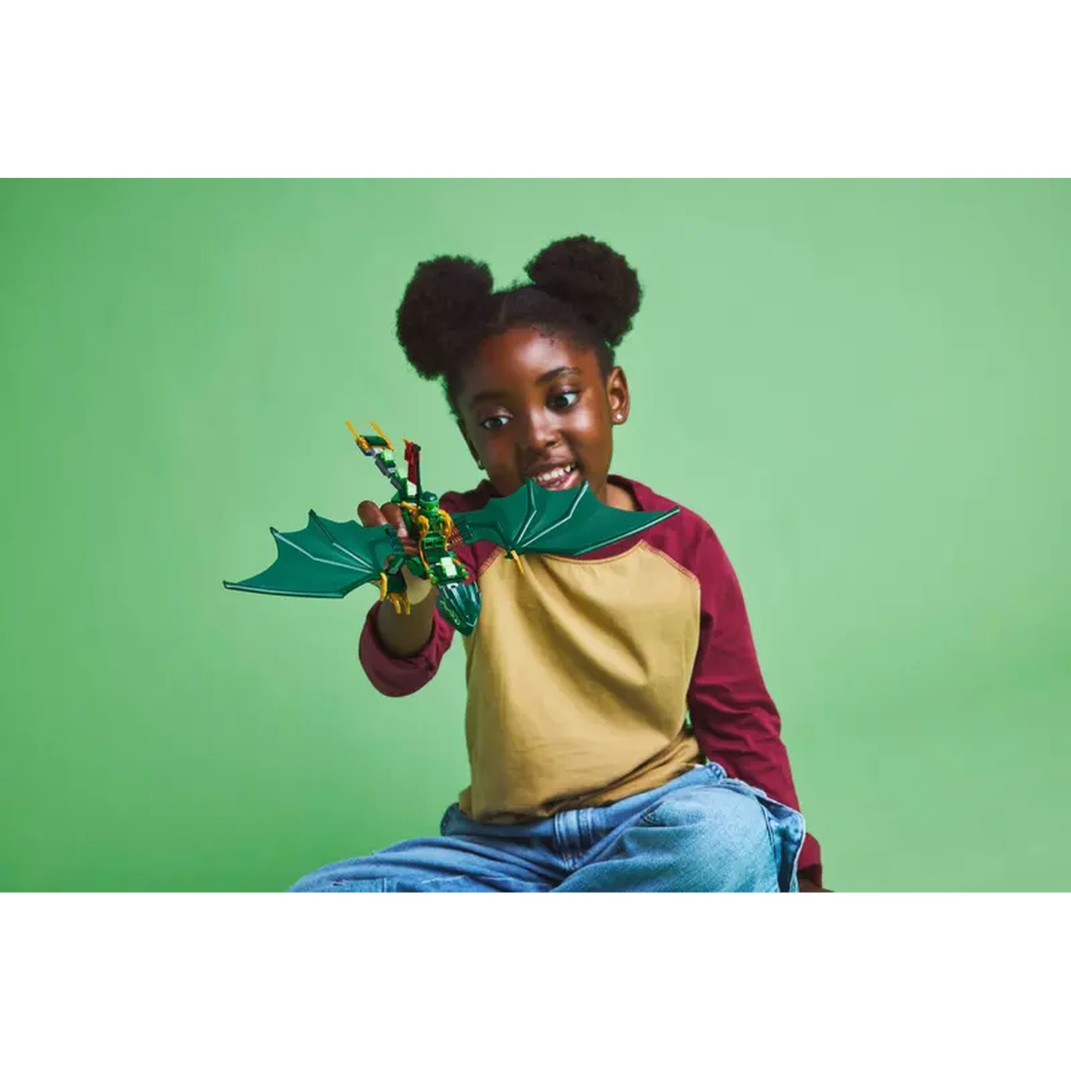 A girl holds a green and gold dragon toy. She wears a maroon and tan shirt and jeans. Green background.