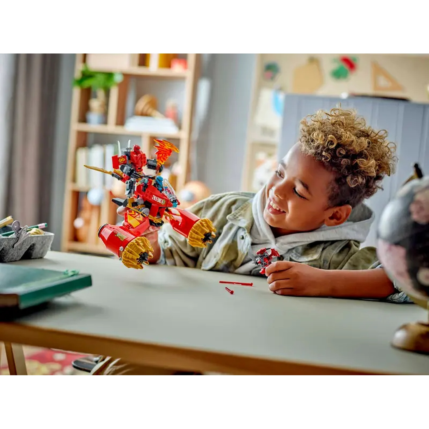 A smiling boy plays with a red Lego Ninjago set, holding a dragon-like vehicle. A globe sits on the right, a desk in foreground.