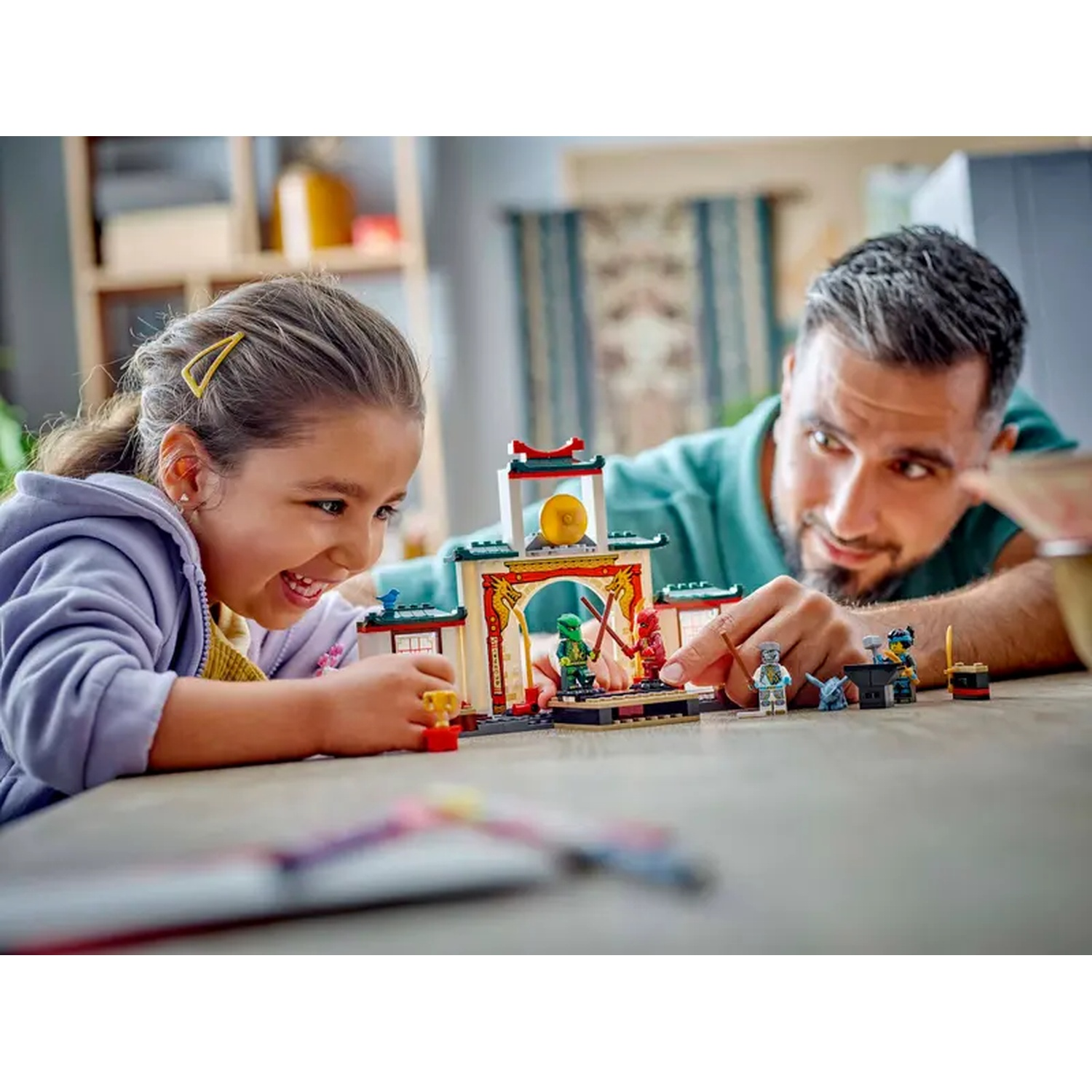 A girl and a man play with Lego. The Lego set features a building with minifigures. Both are smiling and focused on the game.