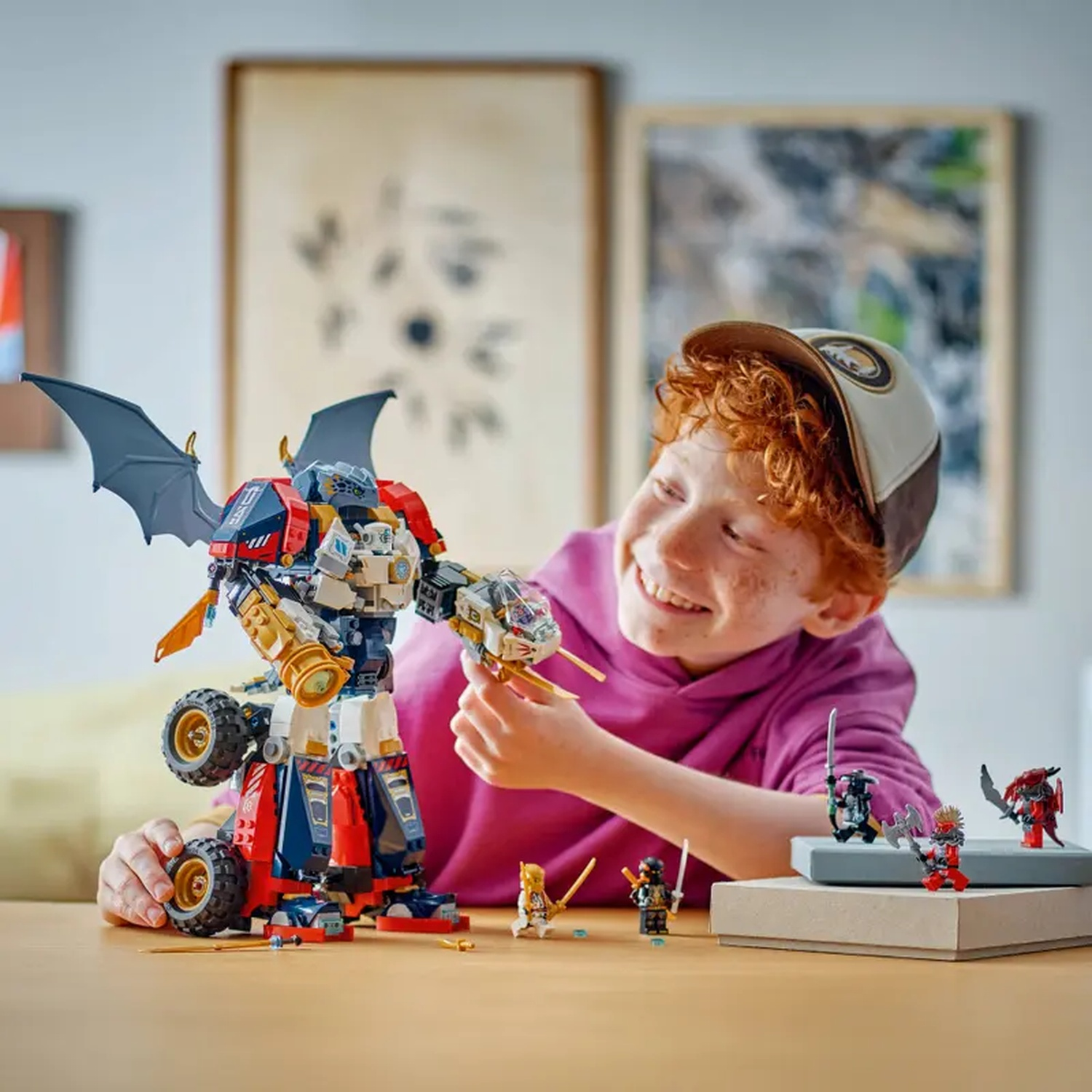Een jongen lacht, speelt met een Lego-robot en mini-figuren op een tafel. De robot heeft wielen en vleugels. Achtergrond: kunst.