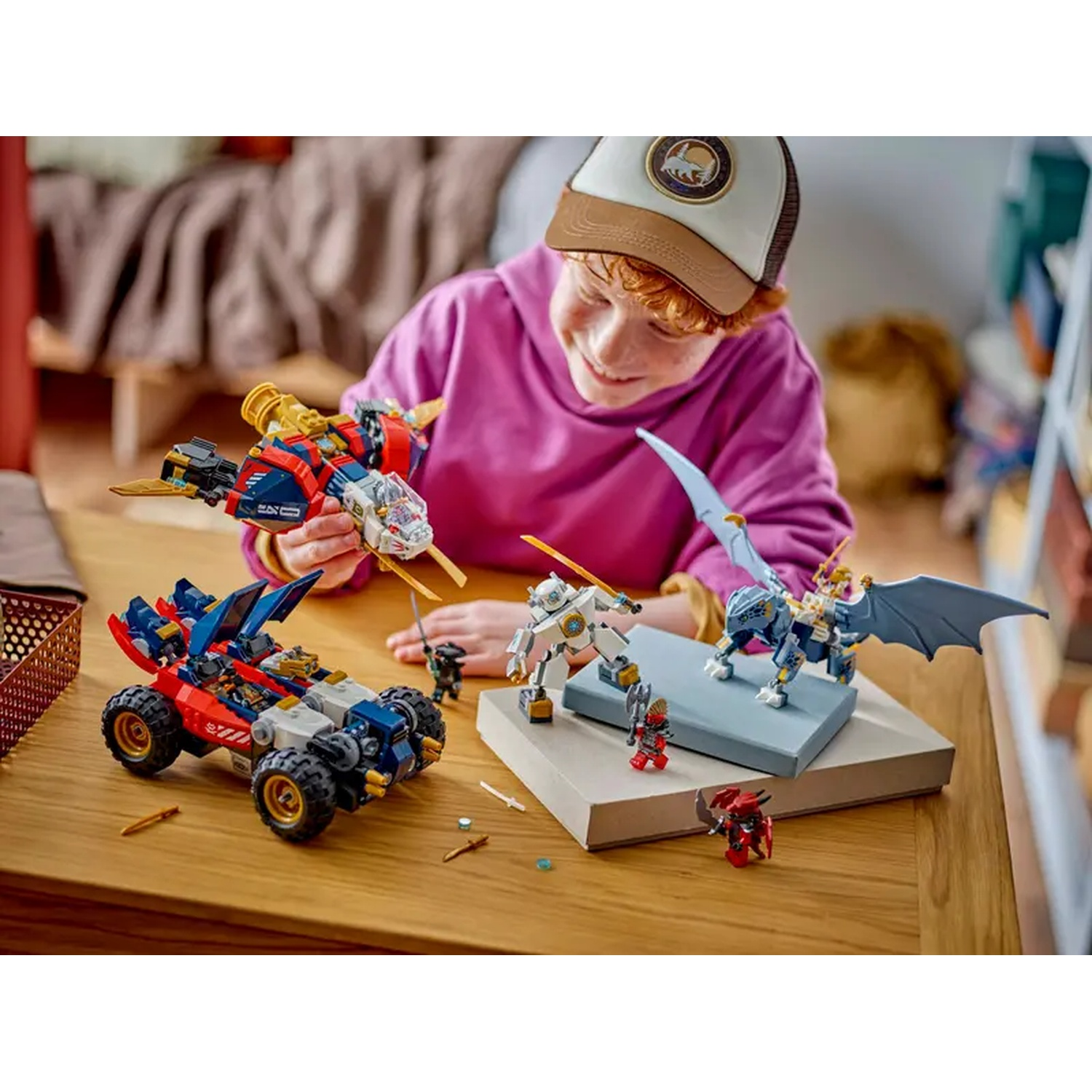 Een kind lacht, spelend met Lego-speelgoed: een voertuig, een straaljager, robots en een draak op een houten tafel. Achtergrond is een slaapkamer.
