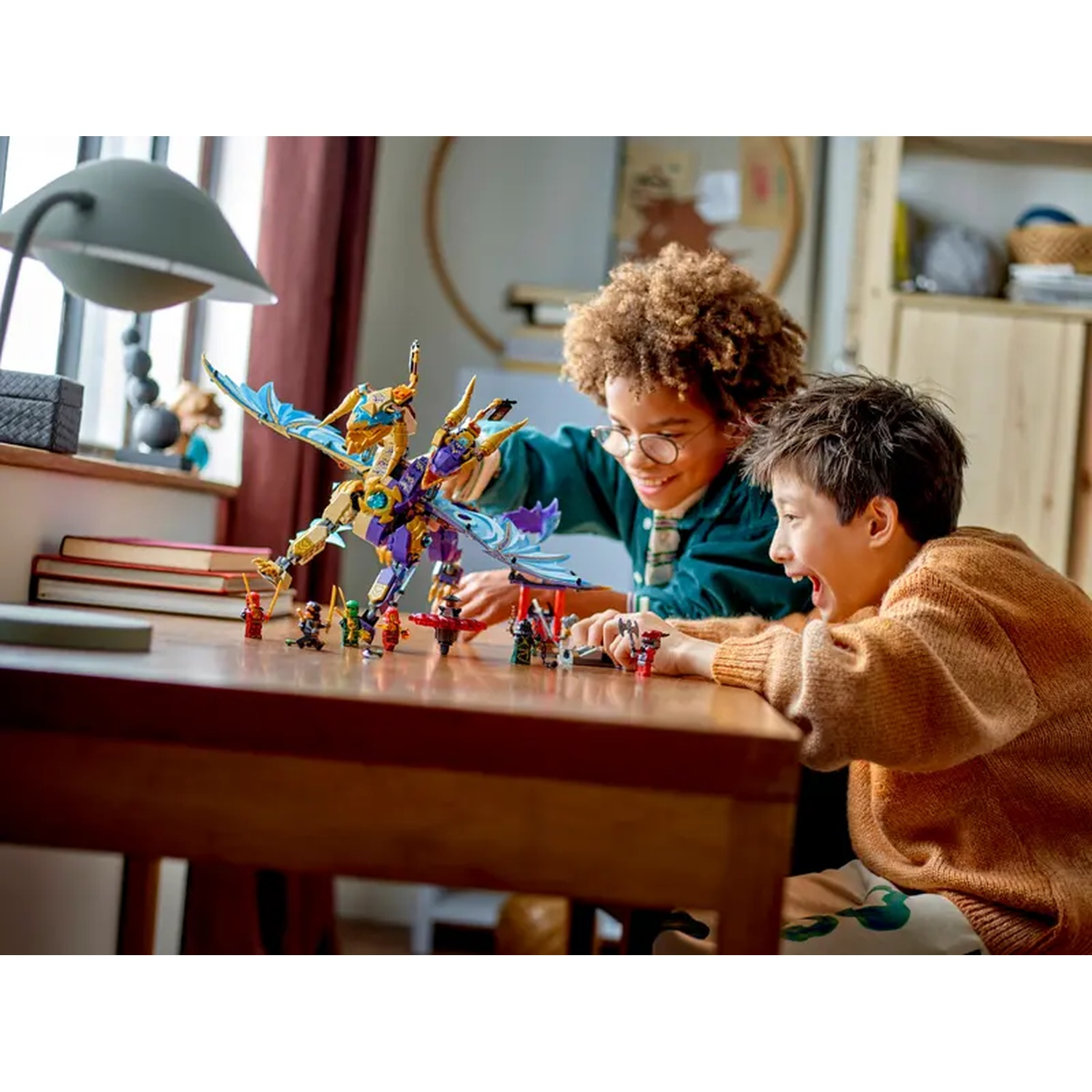 Two boys play with a Lego dragon and figurines on a table. The scene is indoors, with a window and a lamp visible.