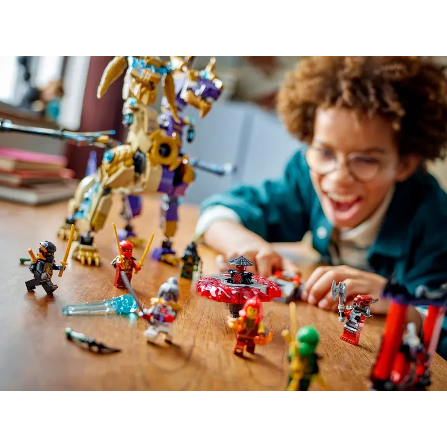 A boy with glasses smiles at a Lego scene. A golden dragon and ninja minifigures are on a wooden table.