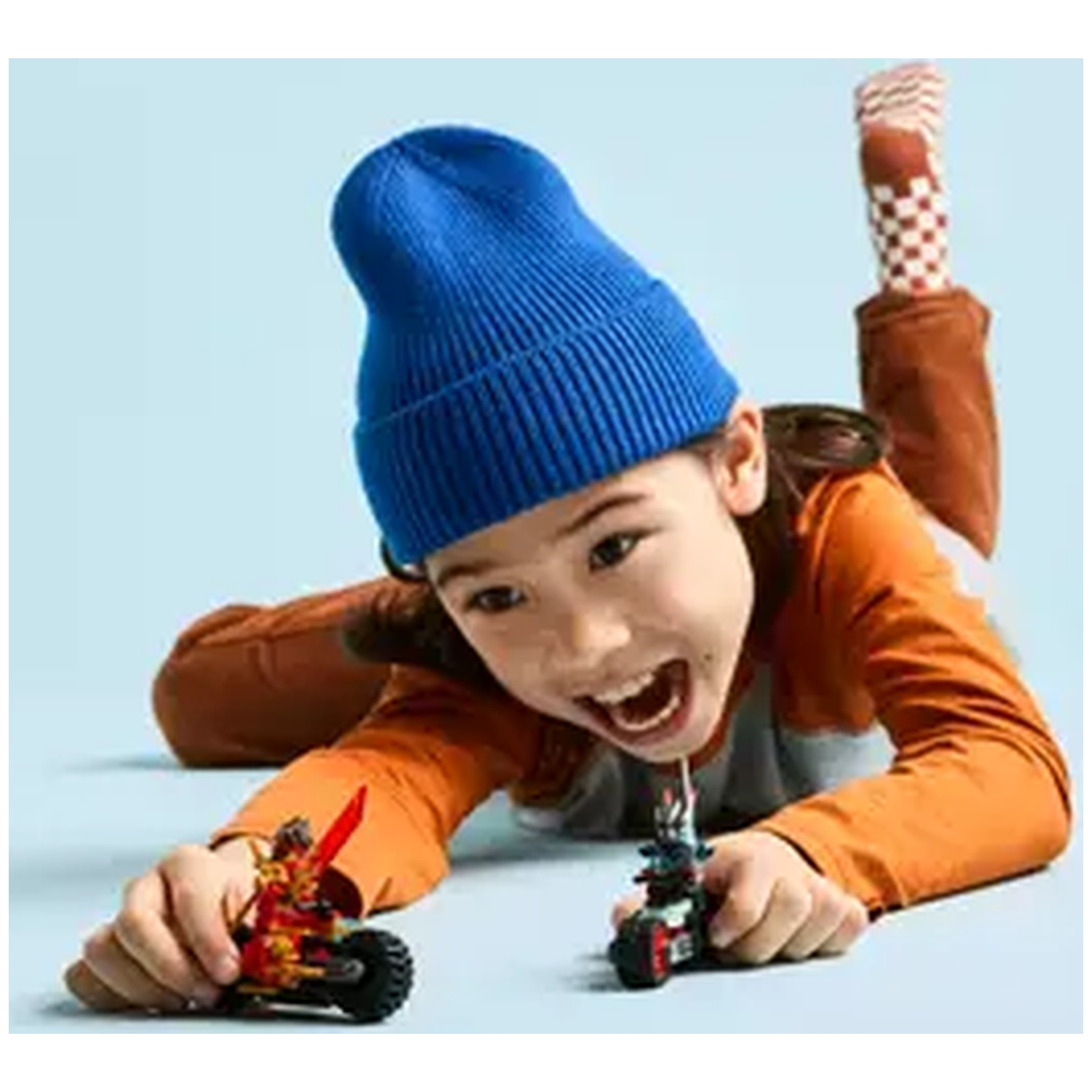 A child in a blue hat and orange shirt plays with toy motorcycles, smiling. Background is light blue.