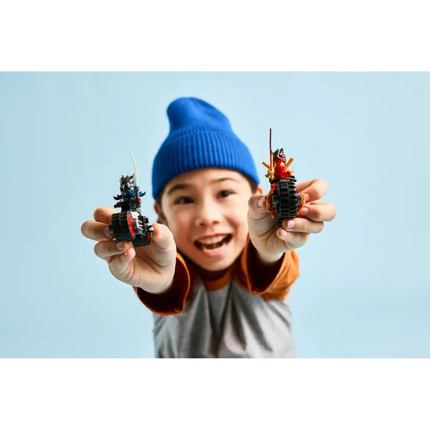 A smiling boy in blue hat holds two toy motorcycles, one red, one black, with a light blue background.