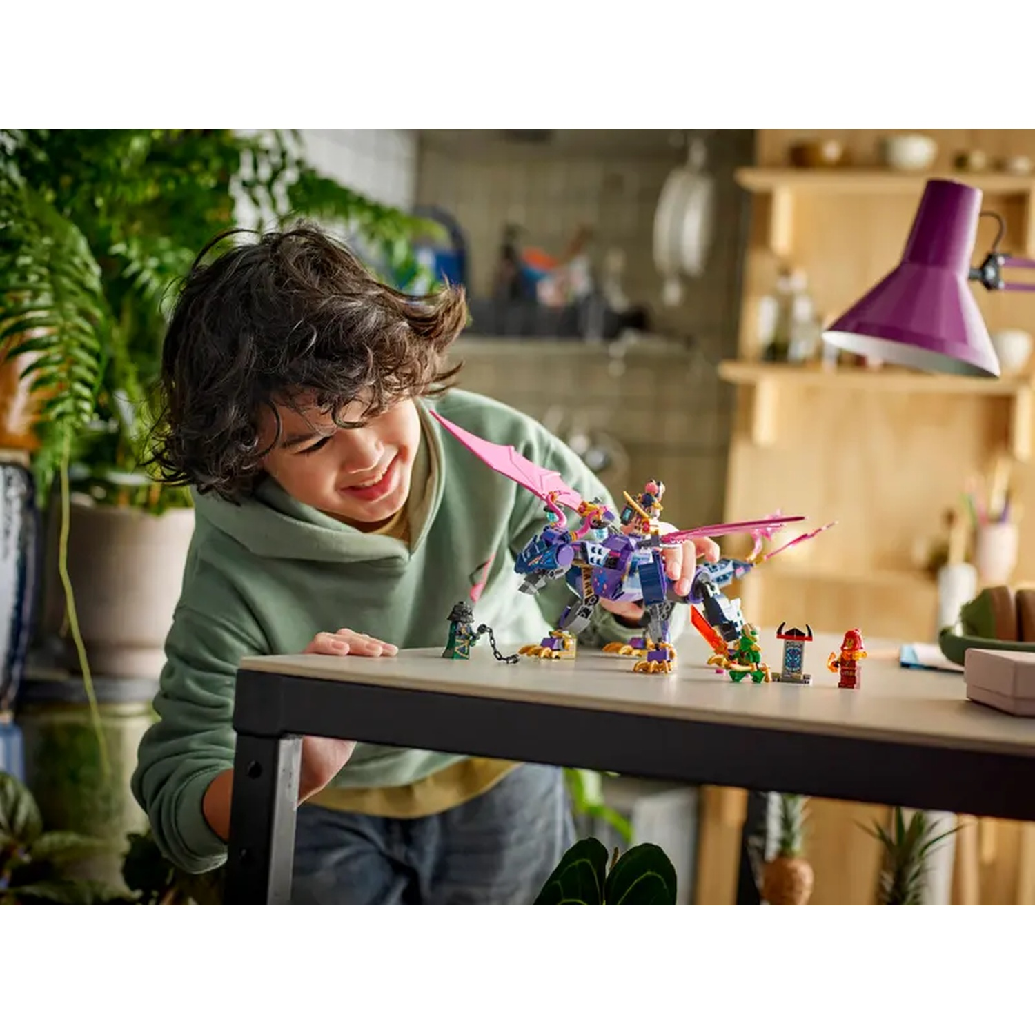 Een kind lacht naar een Lego-draak op een tafel. De draak is paars en roze, met kleine Lego-figuren in de buurt. Een lamp staat op de achtergrond.
