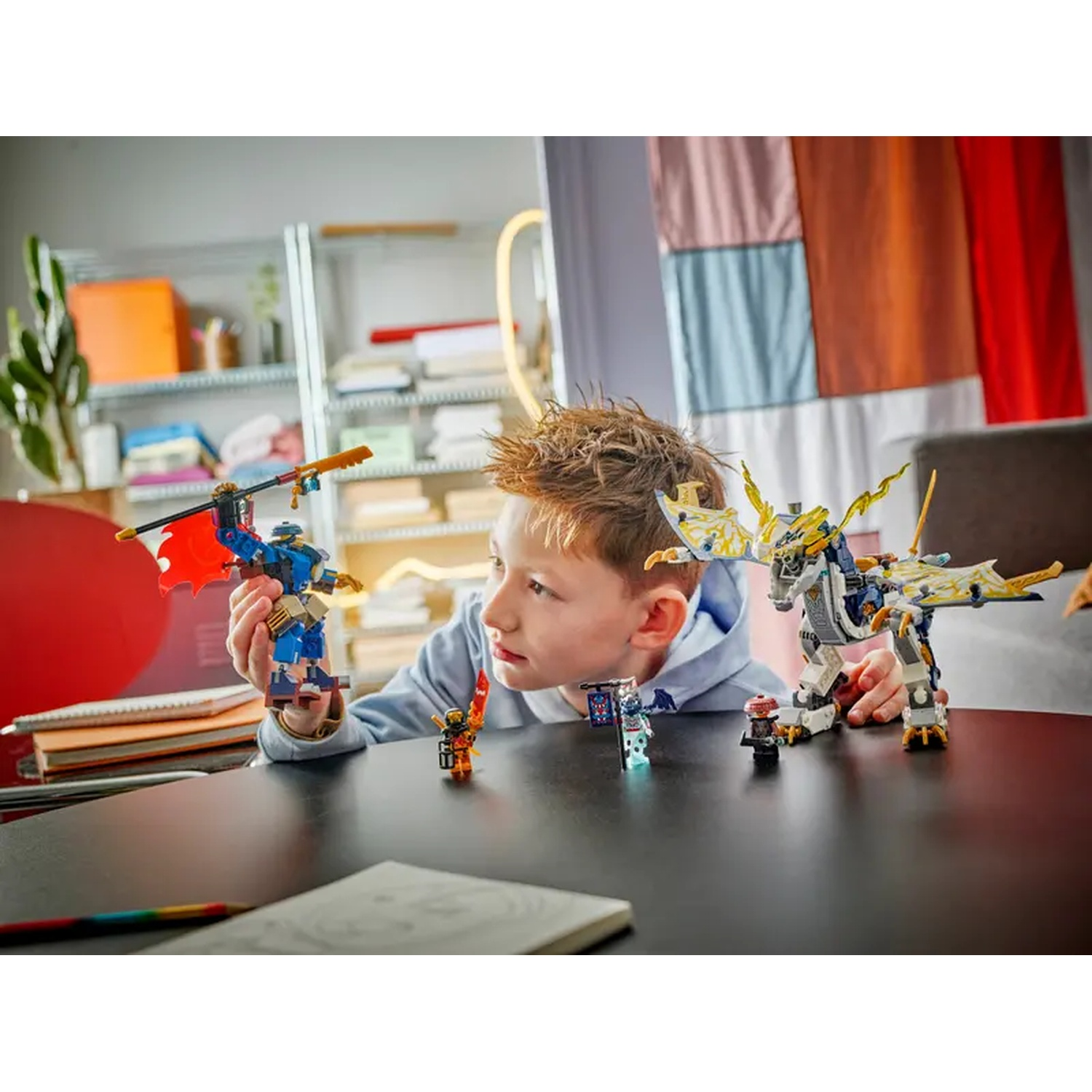 A boy looks at Lego figures on a table. Lego dragon, ninja, and other figures are on the dark surface. Books and a shelf are in the back.