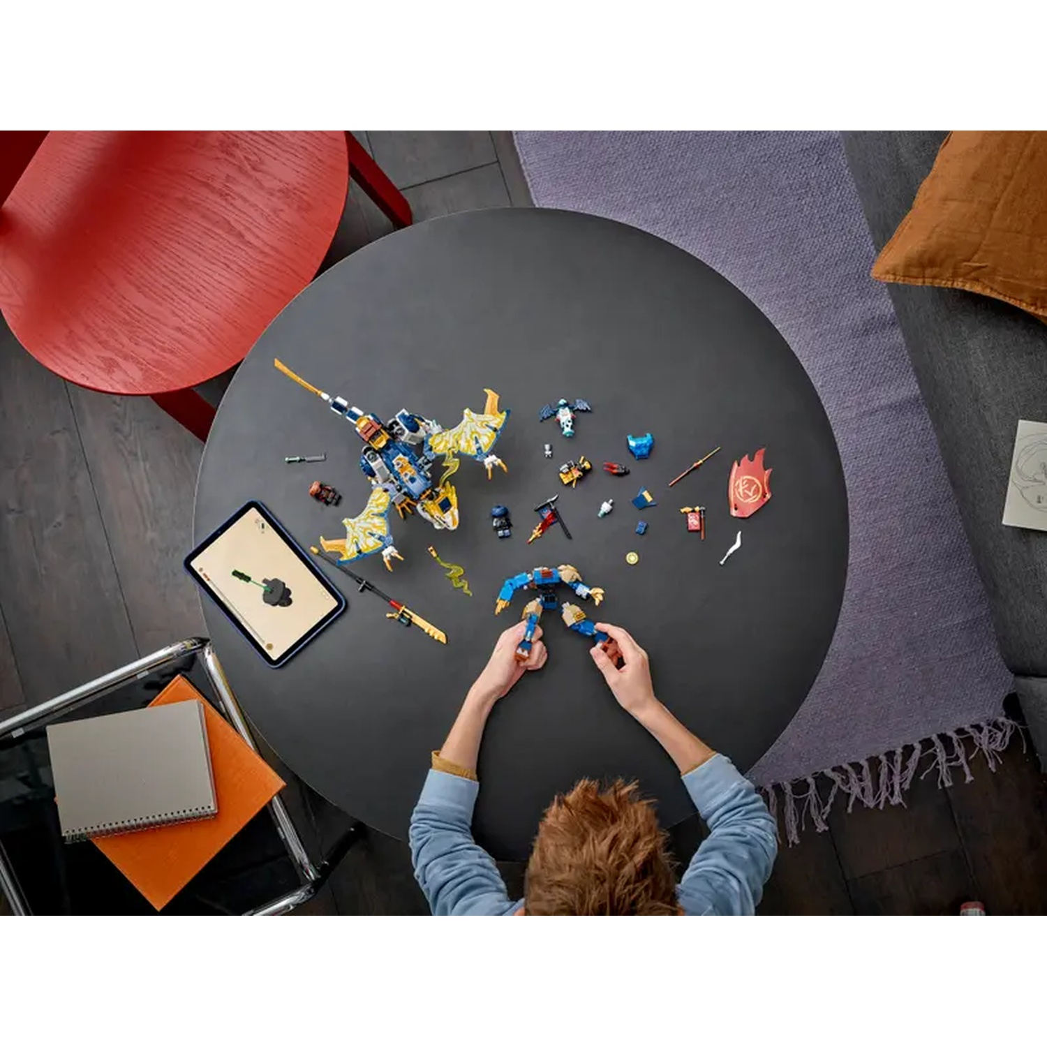 A child is assembling a Lego set on a dark round table. Various Lego pieces are scattered around, along with a tablet.