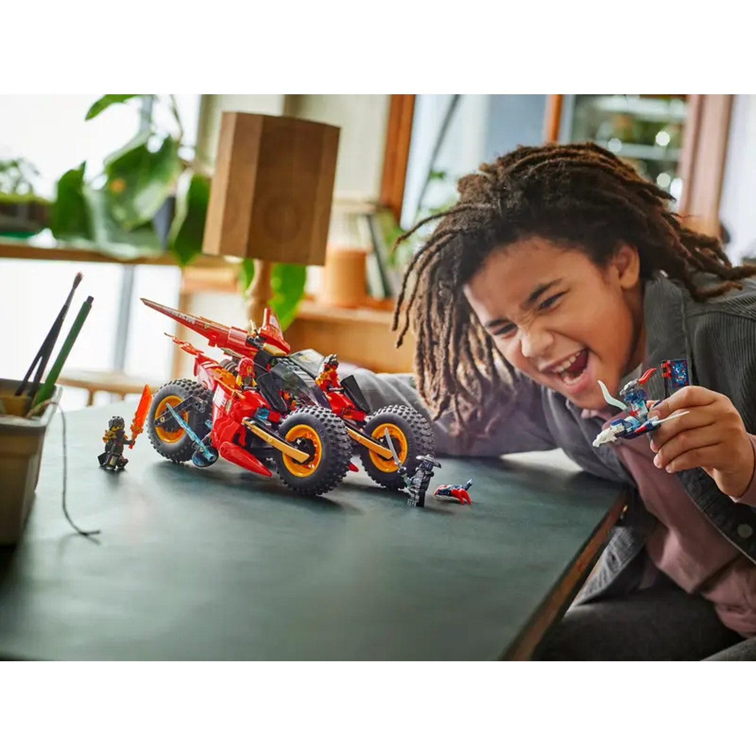 A child smiles holding Lego toys. Red and black Lego car on a green table. Other Lego figures are scattered around.
