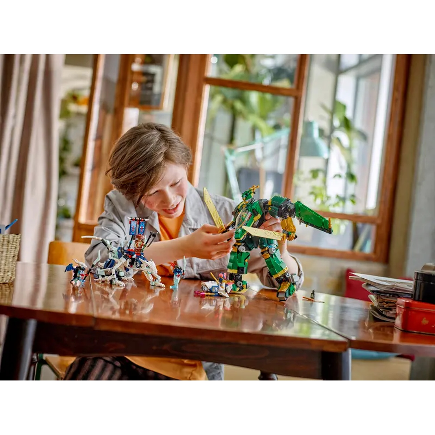 A boy smiles, playing with Lego robots on a table. The robots are green and white. Windows and a curtain are in the background.