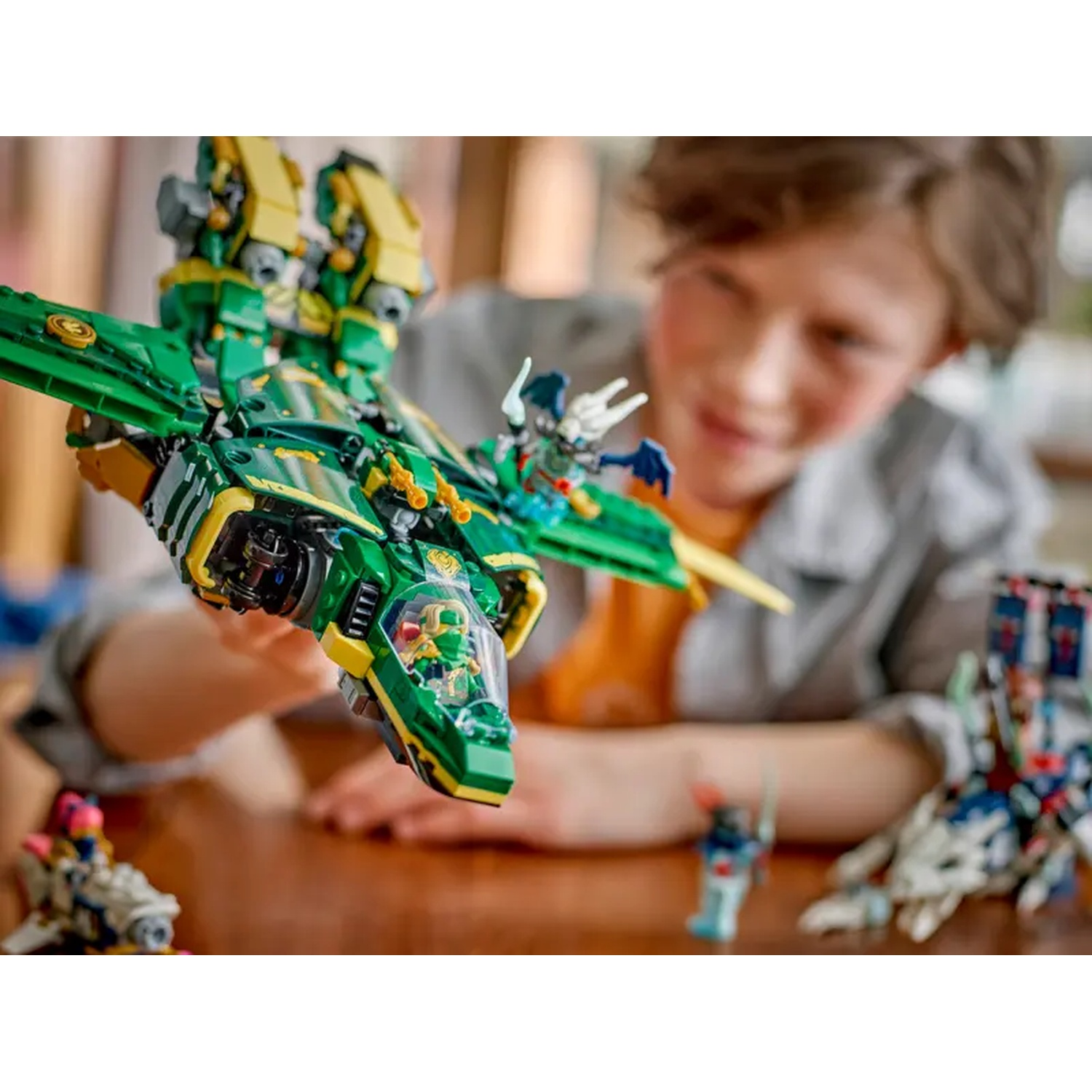 A boy holds a green and yellow Lego jet. Other Lego figures and parts are on the table. The boy is smiling.