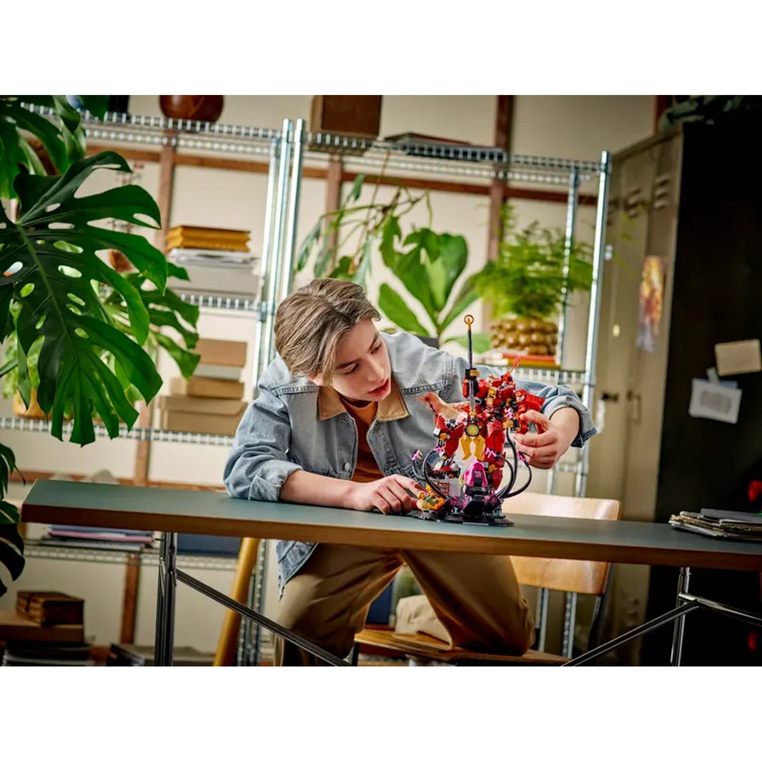 A person is building a red and yellow Lego set on a table. The background includes shelves with plants and a dark cabinet.