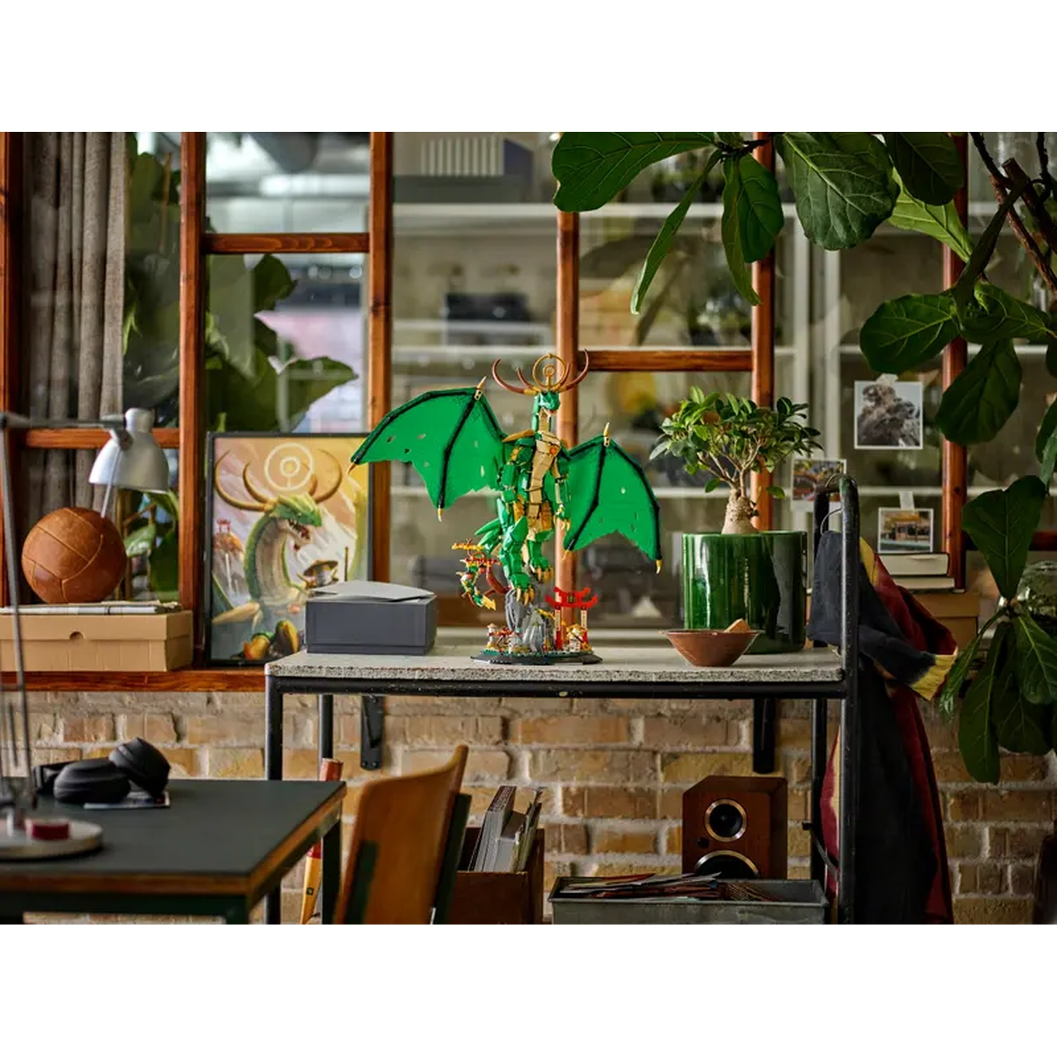 Un dragon Lego sur une étagère avec une plante verte, une peinture et un bureau. L'arrière-plan présente des fenêtres et un mur de briques.