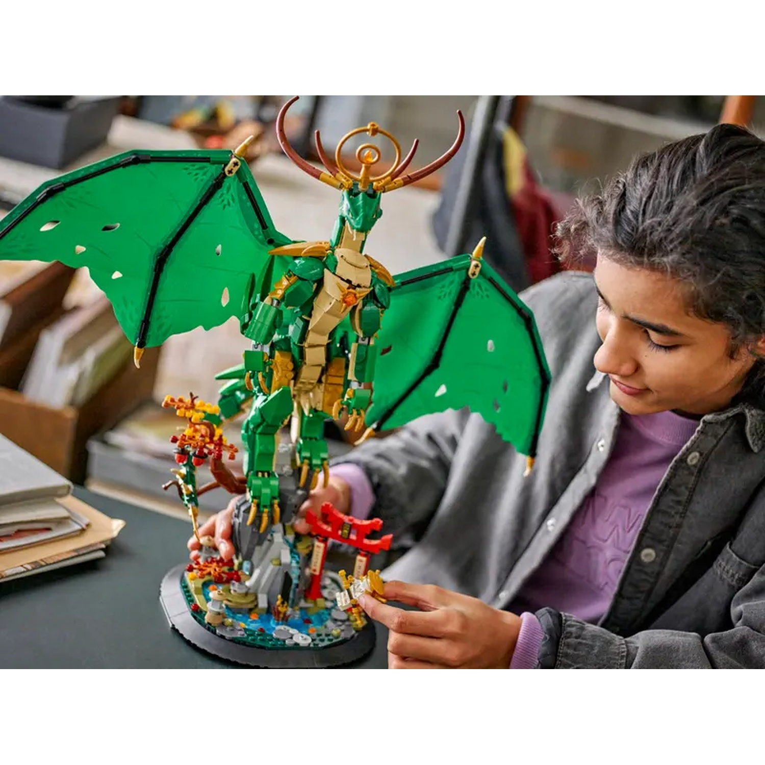 Une personne construit un dragon Lego. Le dragon est vert et doré. La personne porte une veste grise et une chemise violette.