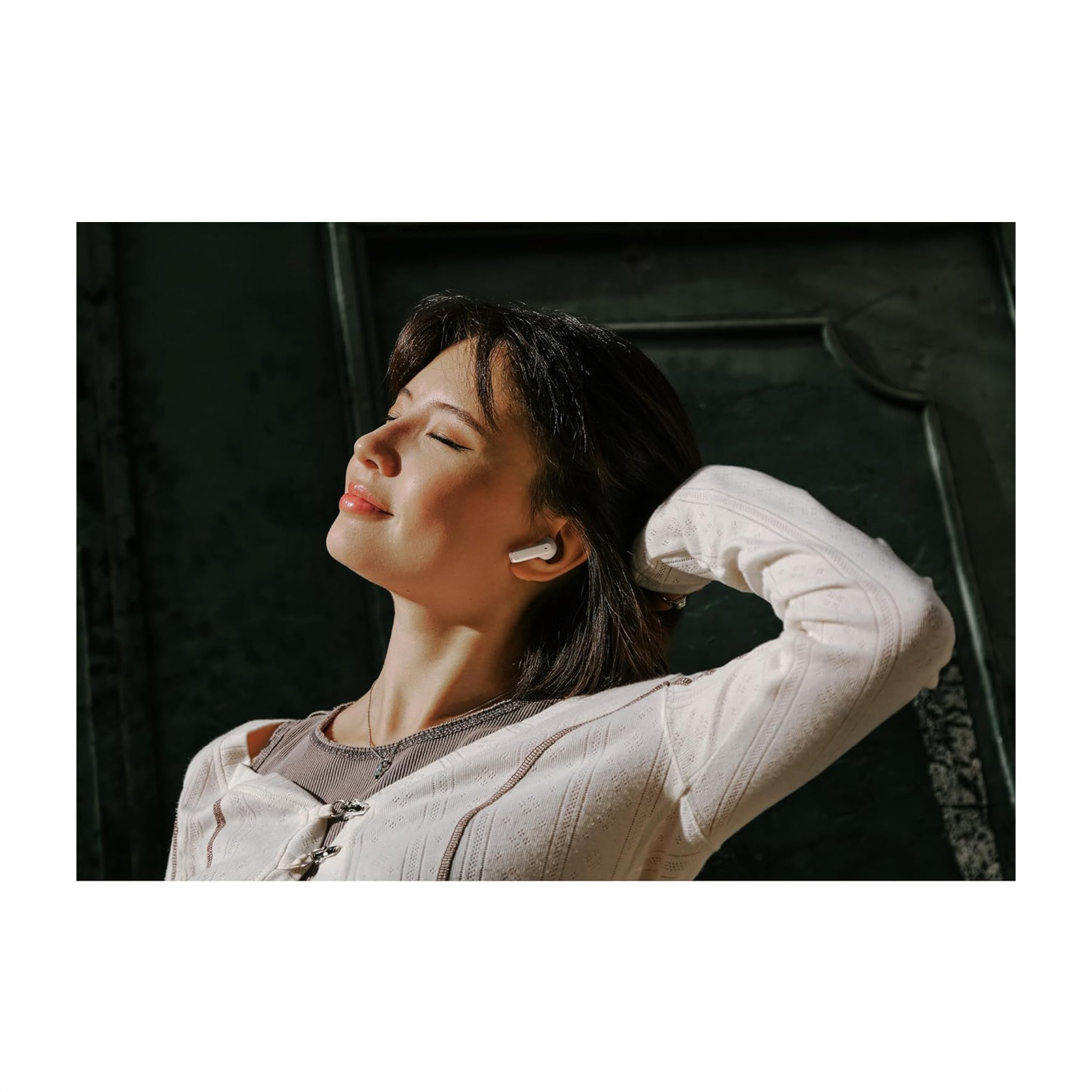 Una donna con un cardigan bianco e auricolari, occhi chiusi, mano che tocca i capelli. Lo sfondo è una porta verde scuro.