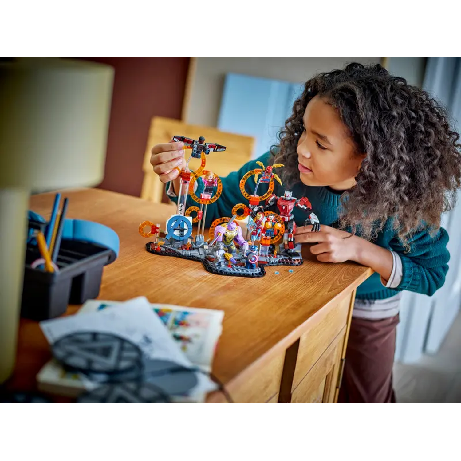 A girl looks at Lego sets on a wooden table. The sets feature colorful characters and structures. A lamp and papers are nearby.