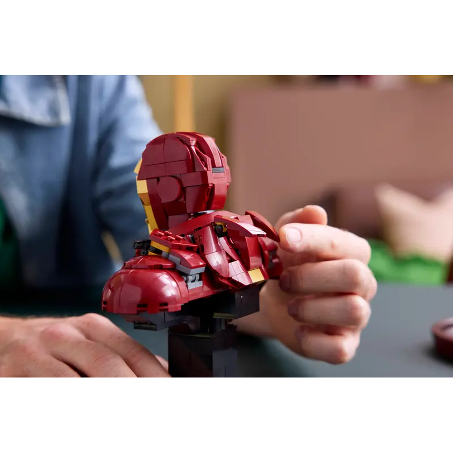 A Lego Iron Man bust, red and gold, on a black stand, held by hands. A person in a blue shirt is in the background.