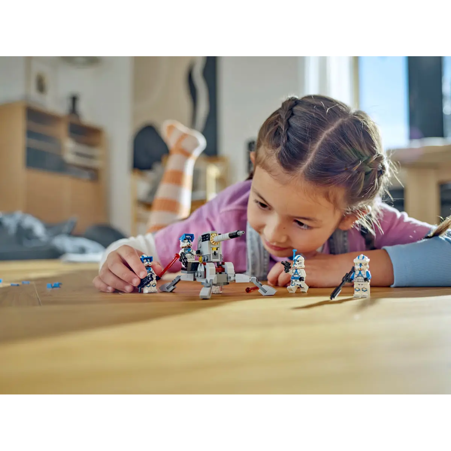 A girl plays with Lego Star Wars figures on a wooden floor. A Lego vehicle and figures are in front of her.