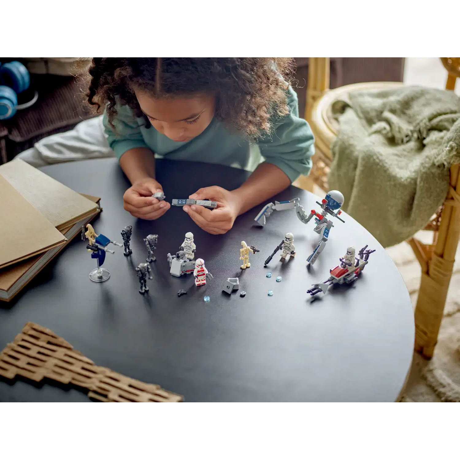 A child is playing with Star Wars Lego on a table. Lego figures are spread around. Background shows a chair and blanket.