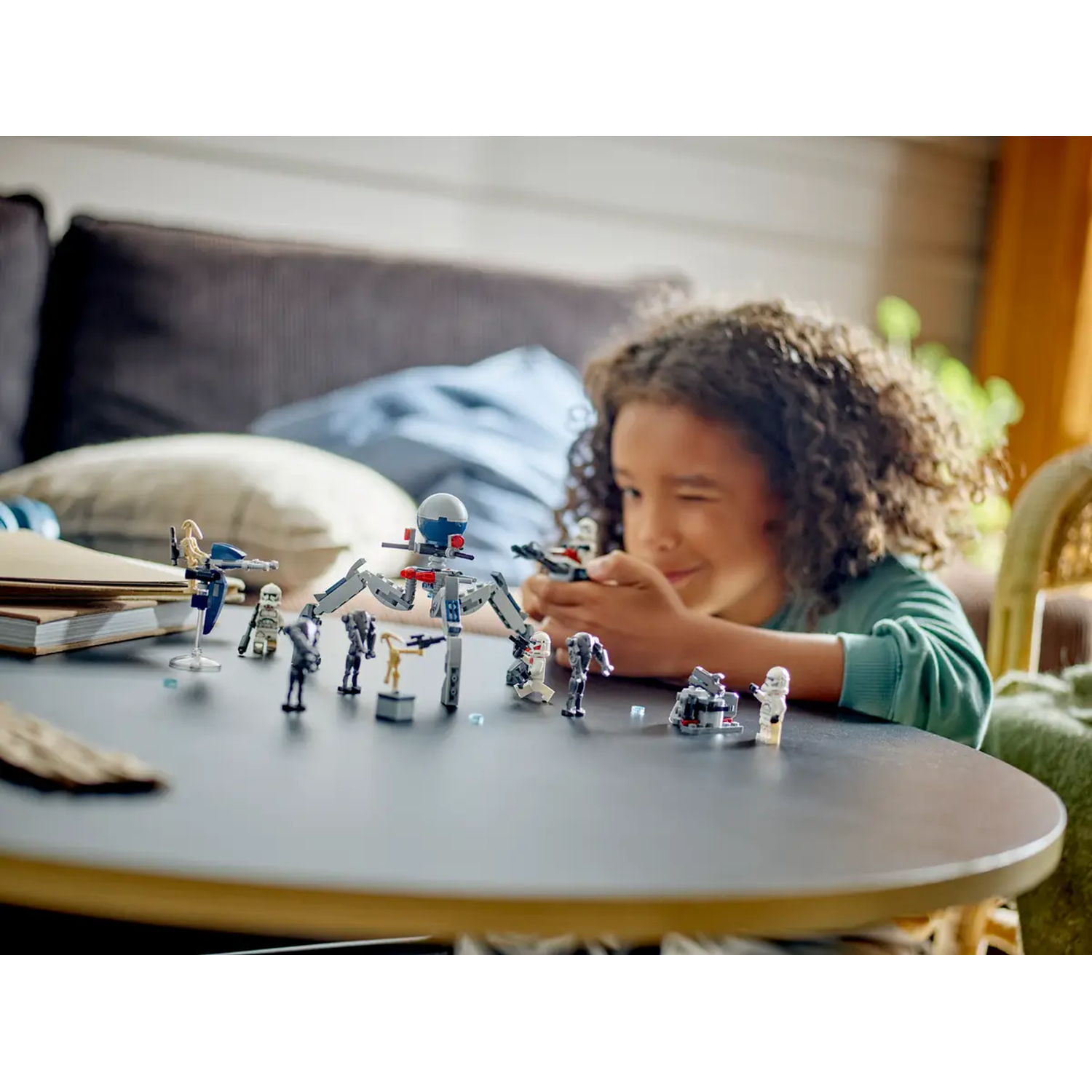 A child is playing with Star Wars Lego on a table. Lego figures and a mechanical spider are on the table. A couch is in the background.