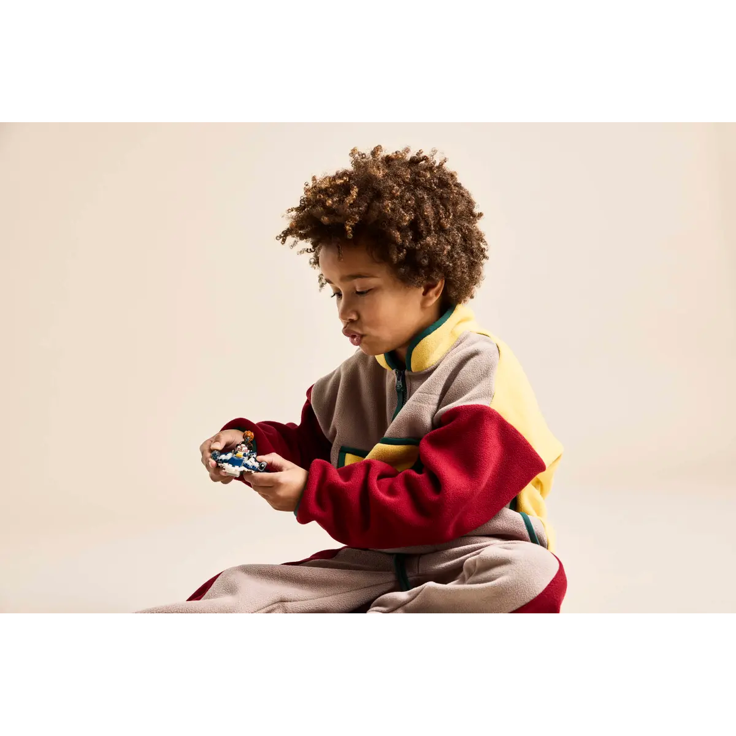 A child in a color-block outfit holds a small toy. The outfit is gray, yellow, and red. The background is a cream color.