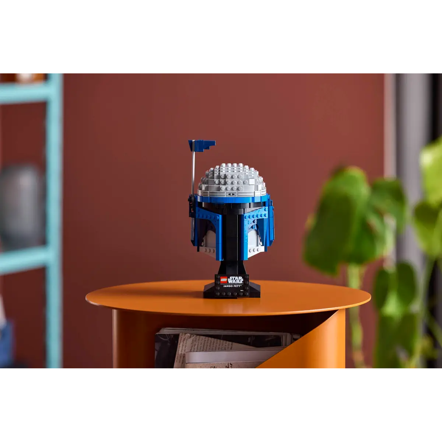 A Lego Star Wars Jango Fett helmet sits on an orange table. A teal shelf and a green plant are in the background.