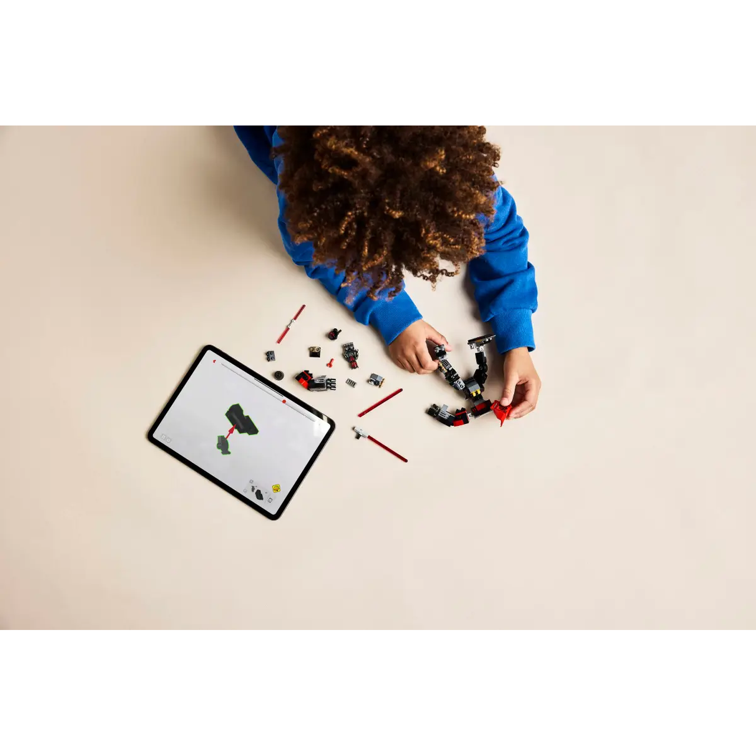A child in blue assembles a Lego set. A tablet displays instructions. Lego pieces, red sticks, and a partially built structure are visible.