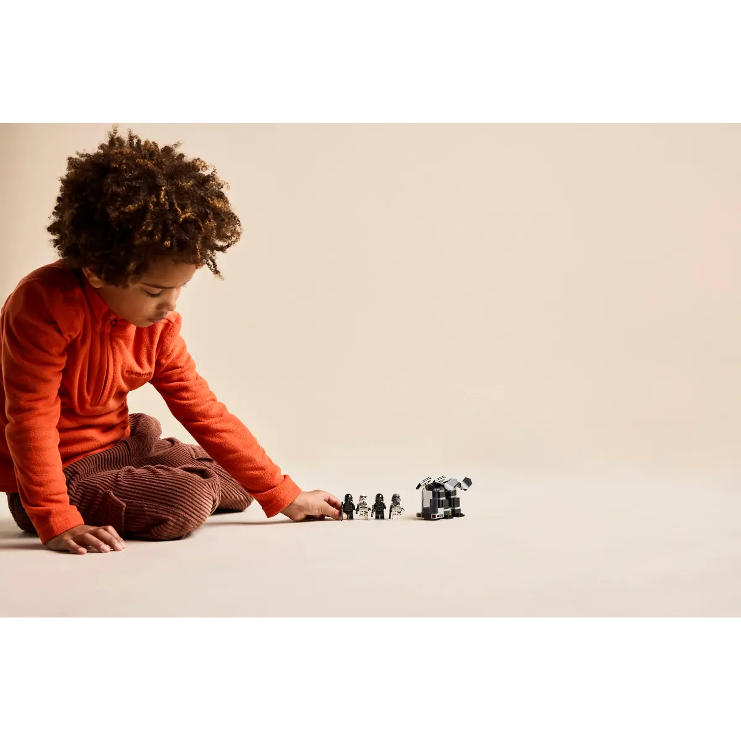 A child in an orange shirt and brown pants plays with Star Wars Lego figurines and a vehicle on a beige surface.