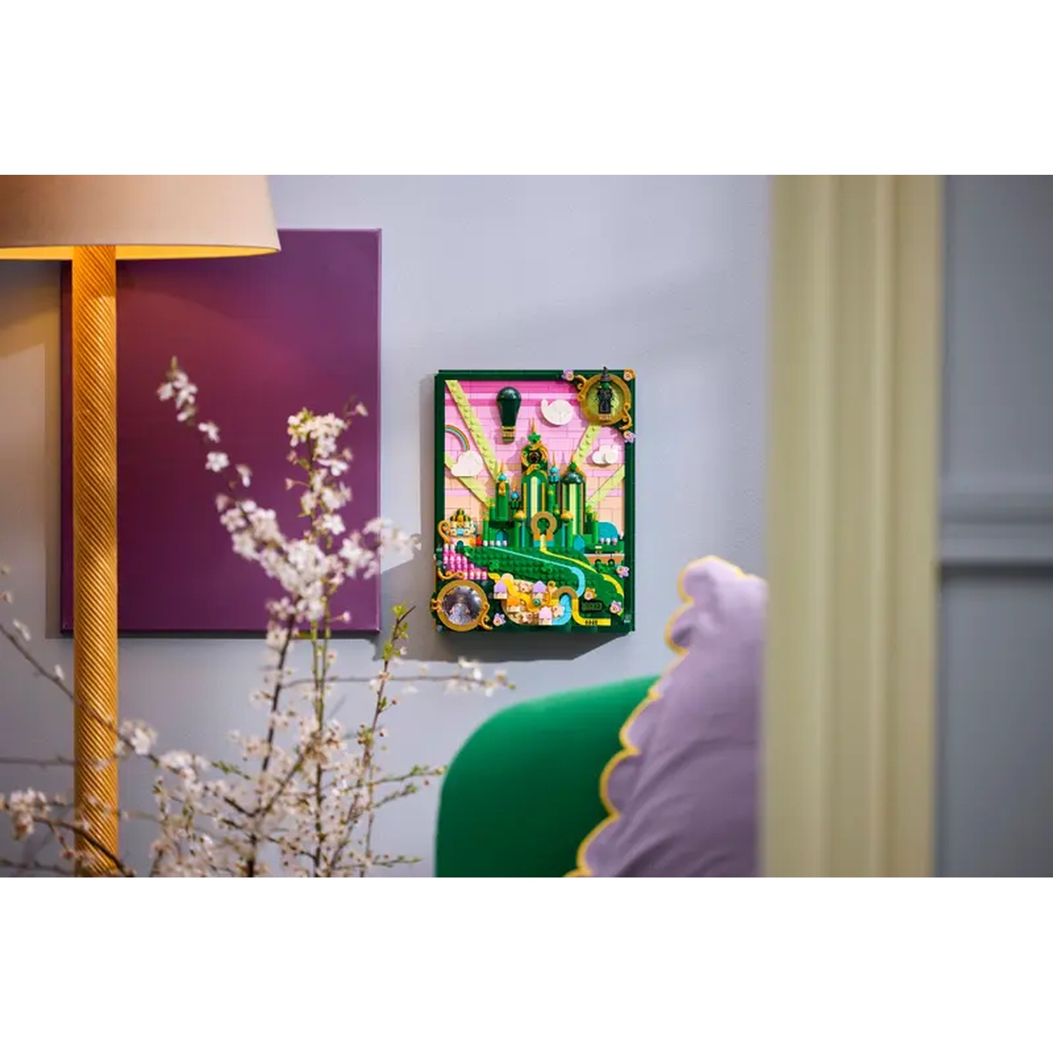 Een ingelijste afbeelding van een groene stad met een gele weg. Een paars schilderij en een lamp links. Een groene stoel rechts.