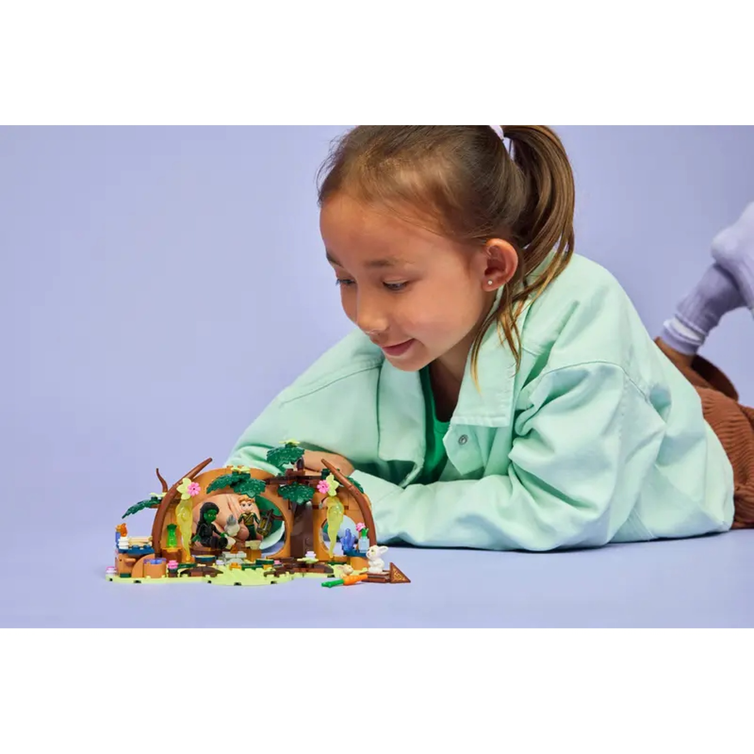 Une fille regarde un jeu Lego représentant une cabane dans un arbre. Elle est allongée sur une surface violette. Le jeu contient des figurines et des plantes.