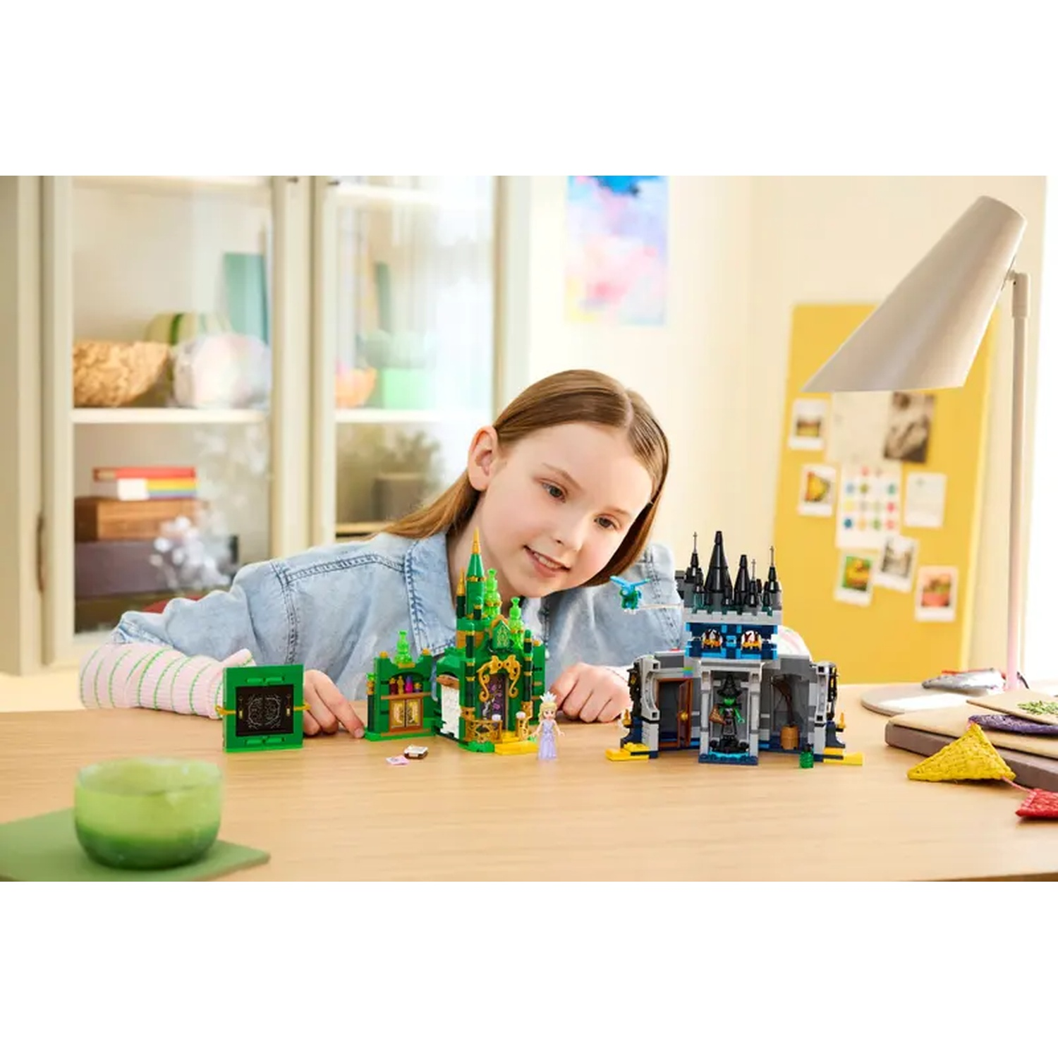Une jeune fille sourit à un château Lego sur une table. Une bougie verte, une lampe et d'autres jouets sont présents.