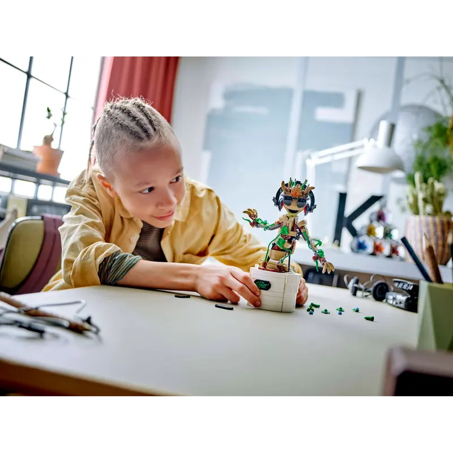 A child is building a Lego figure on a table. The figure is green, brown, and has sunglasses. The child is wearing a yellow jacket.